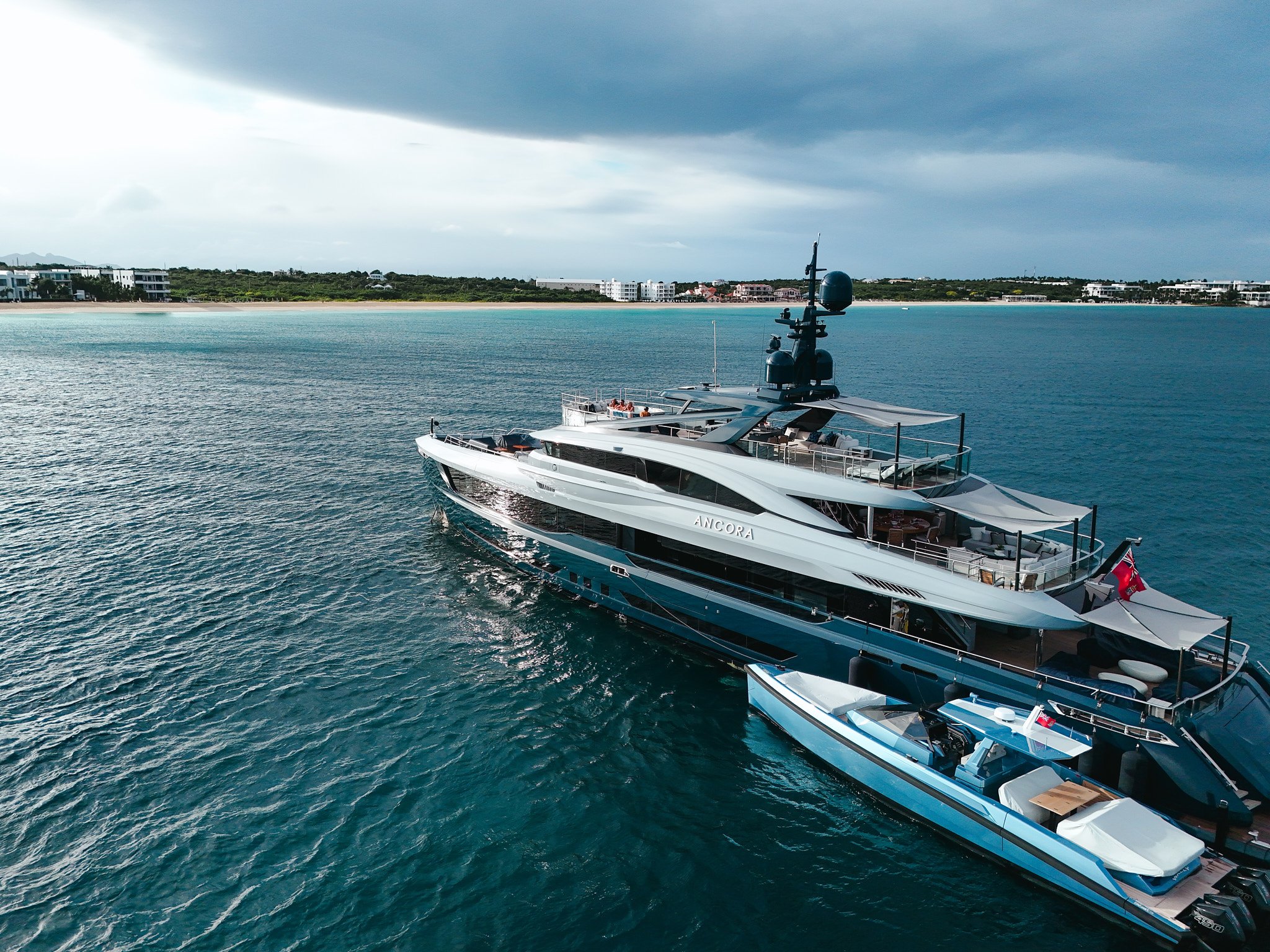 An aerial view of a large luxury yacht named 'Ancora' sailing on the ocean near a coastline with buildings and greenery under a cloudy sky.