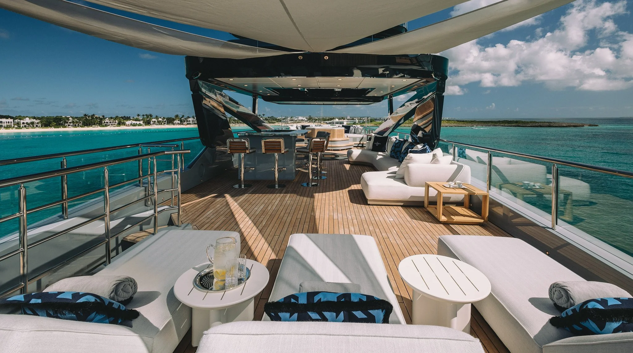 View of the deck on a yacht with white lounge chairs, tables with drinks, a curved sofa with pillows, and a bar area in a tropical location with clear blue water and sky.