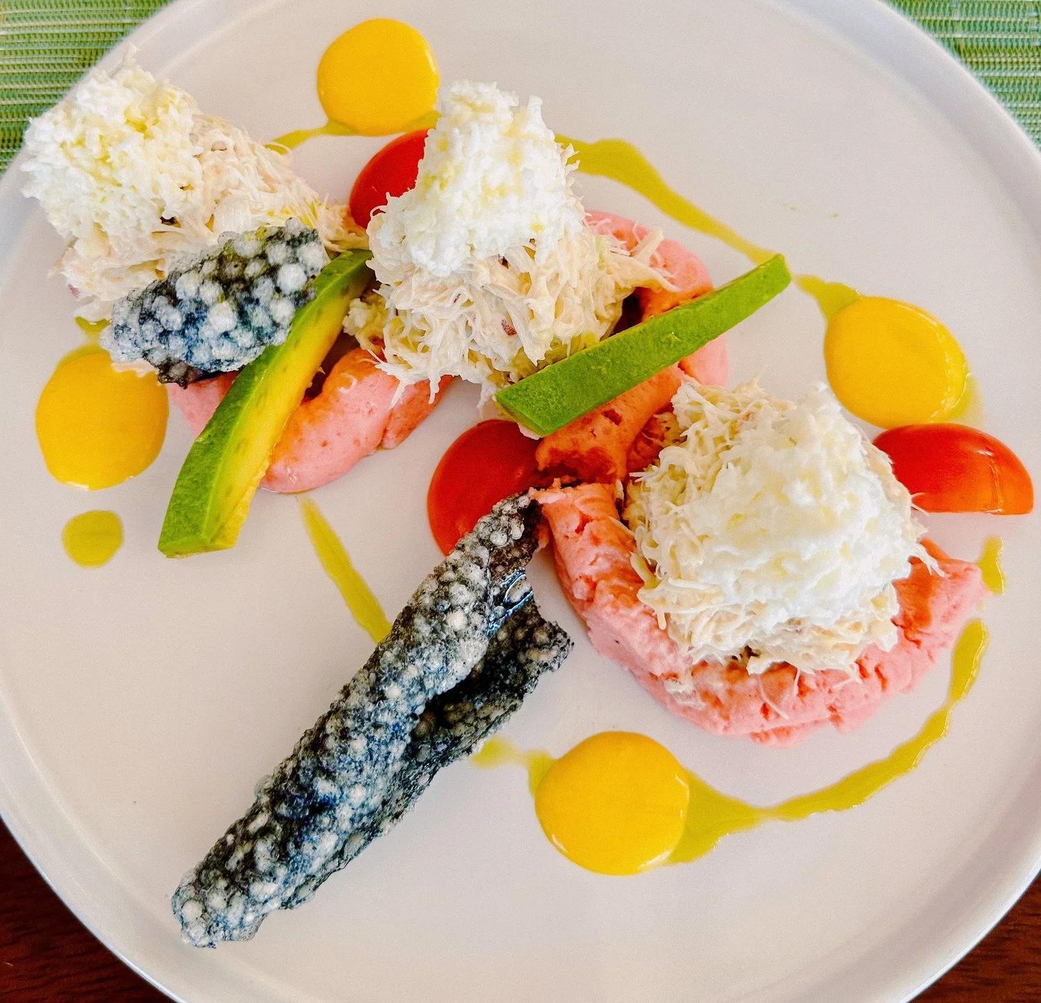 A gourmet seafood dish with two pieces of crab meat topped with shredded cheese and sour cream, garnished with cucumber, cherry tomatoes, and slices of yellow and red sauce dots, served with battered and fried fish fillets