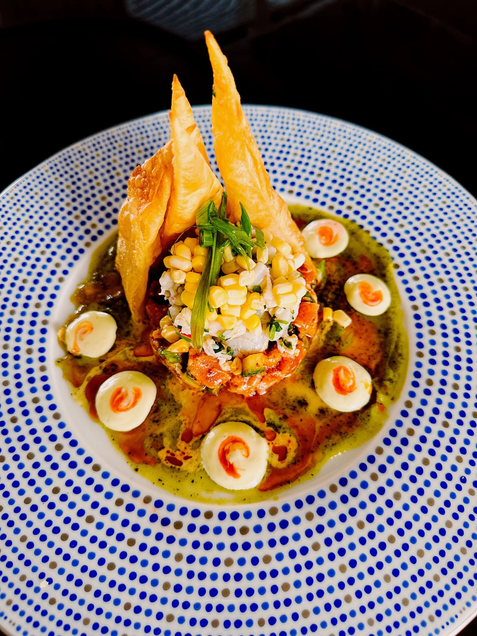 A plated dish featuring a central portion of seafood atop a bed of vegetables, garnished with a sprig of cilantro, surrounded by dollops of mayonnaise with red sauce, and crispy chips standing upright, all on a white plate with blue dots.