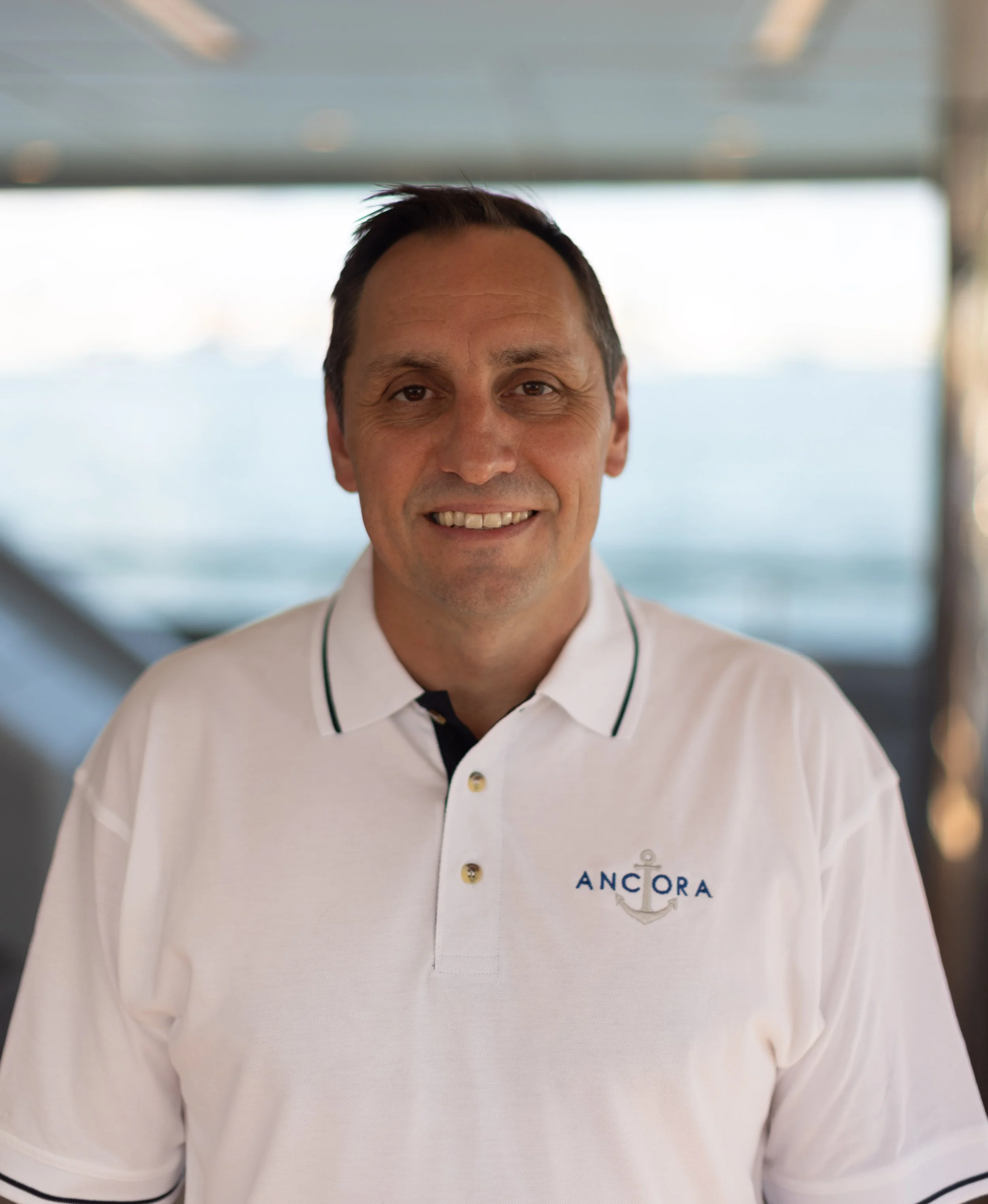 Portrait of a smiling man in a white polo shirt with an anchor logo, standing indoors with a blurred background and ocean view.
