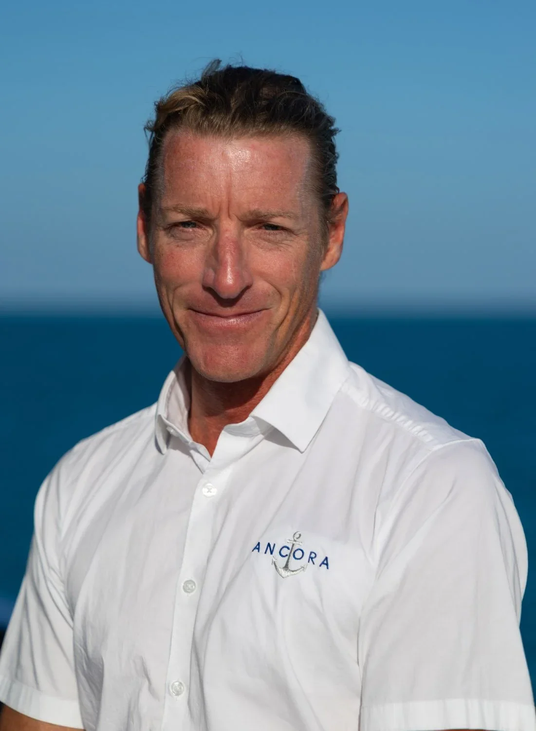 A man with short hair, smiling slightly, wearing a white shirt with the logo 'ANCORA' and an anchor emblem, standing in front of a blue ocean and sky background.
