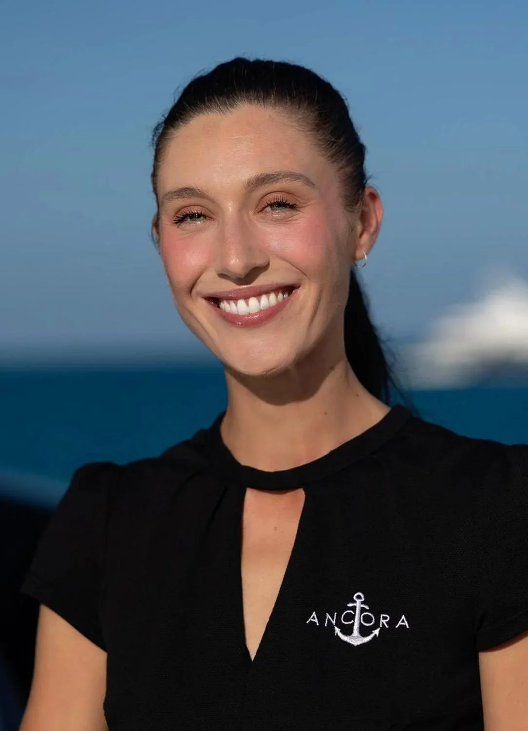 A woman smiling outdoors against an ocean background, wearing a black shirt with a white anchor and 'ANCHORA' logo.