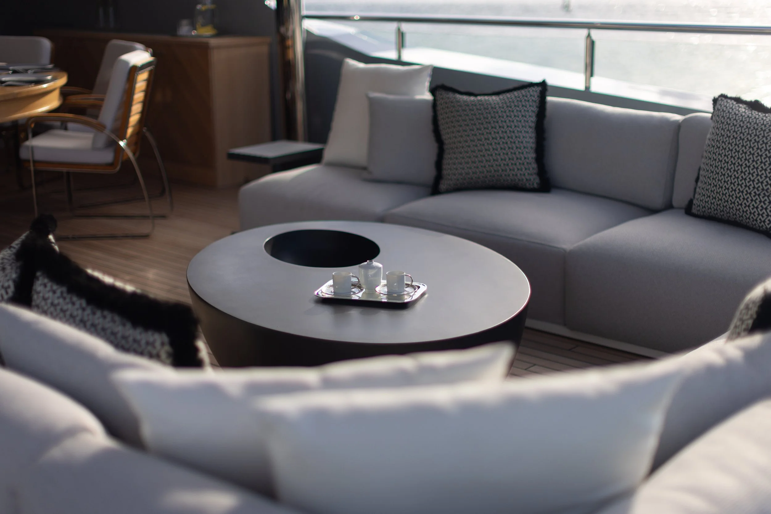 Modern living space with white sectional sofa, patterned throw pillows, a black and white coffee table with three cups and a sugar container, and large windows with sunlight.