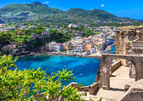 An old brick and stone ruin sits on top of a hill overlooking a villa with a blue marina.
