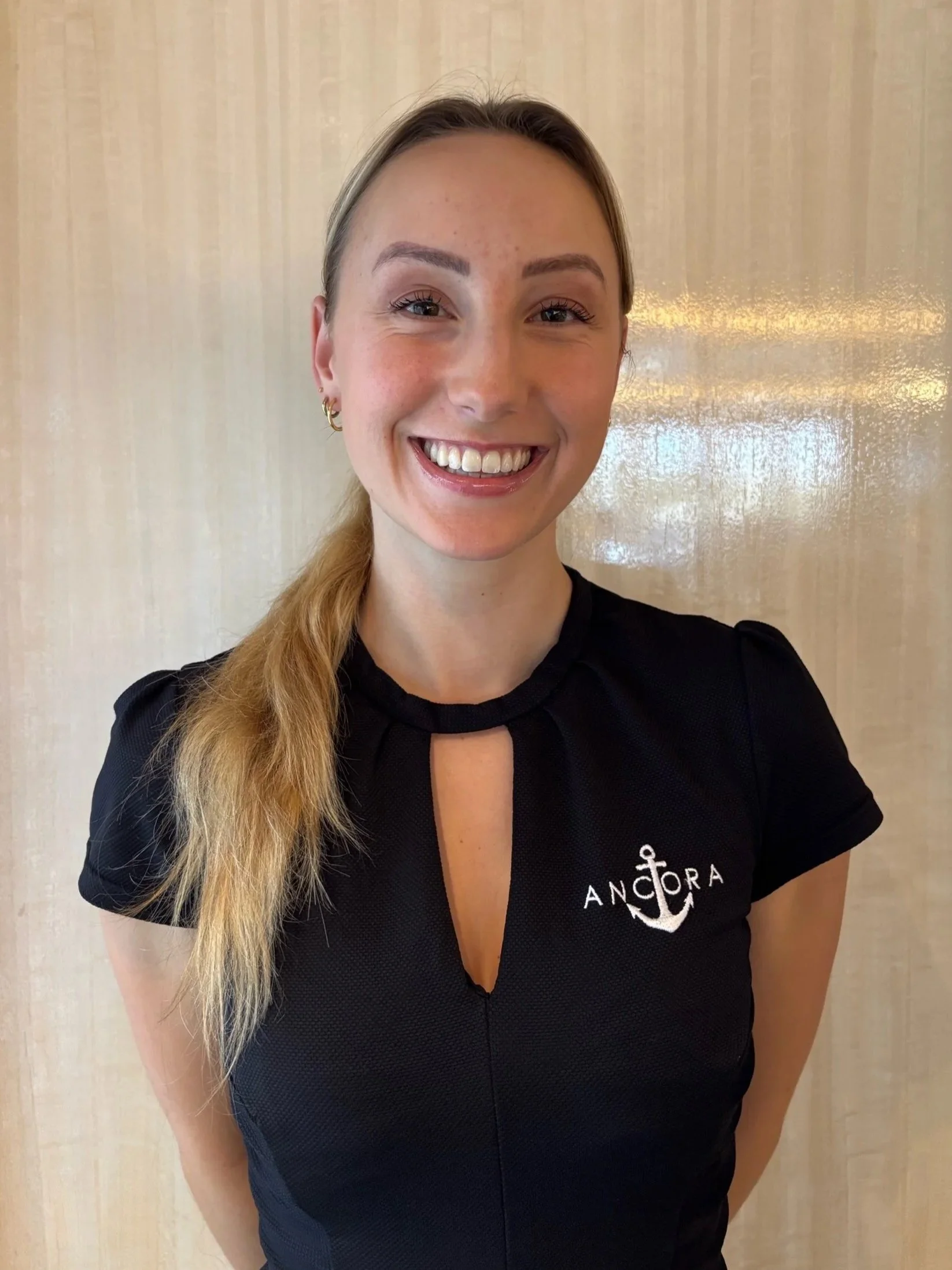 A smiling woman with light skin, long blonde hair in a side ponytail, wearing gold earrings and a black shirt with an anchor logo and the word 'ANCHORA' on it, standing in front of a beige wooden background.