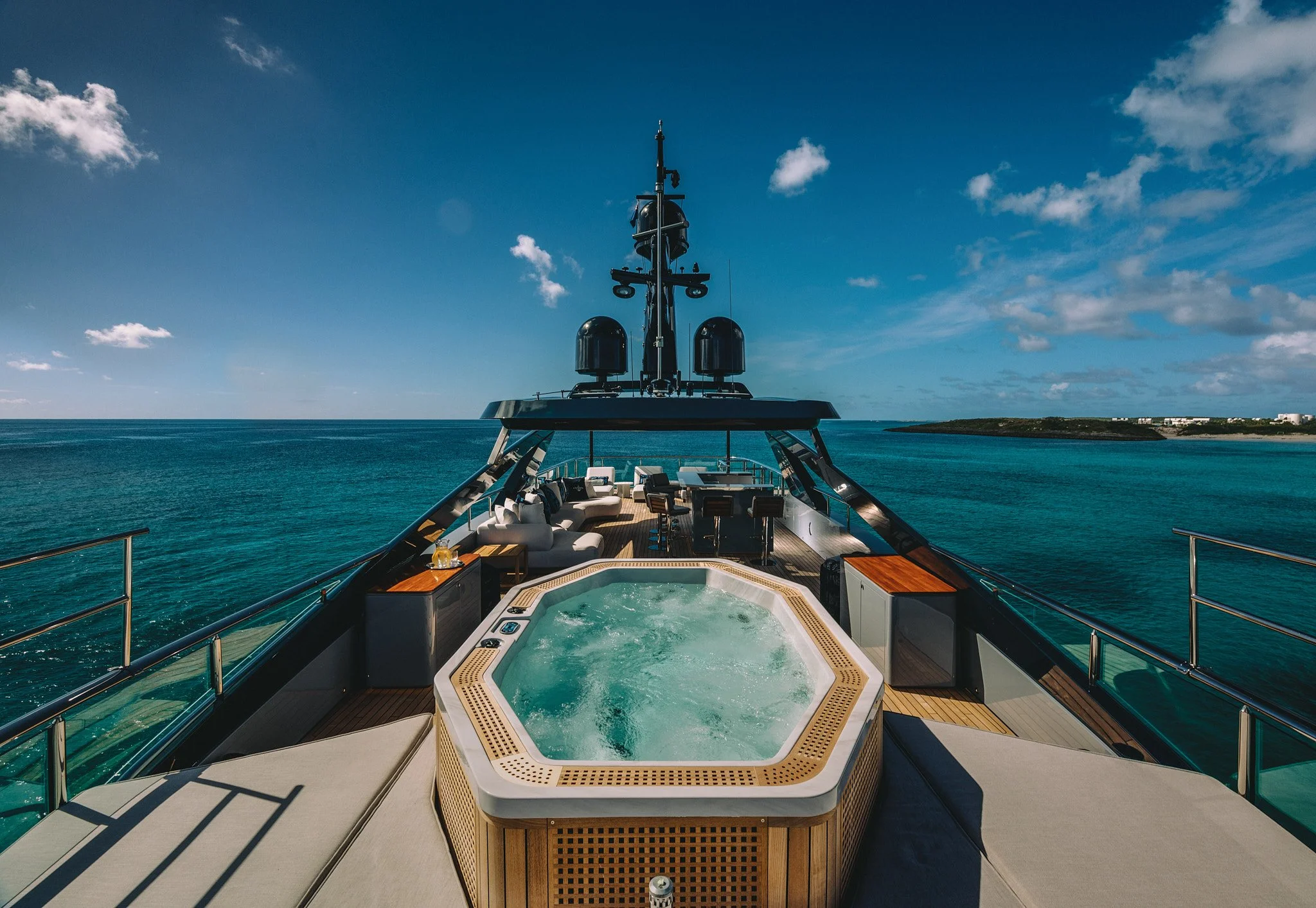 On the deck of a luxury yacht, there is a hot tub filled with water, surrounded by outdoor seating and tables, with a view of the ocean and a distant shoreline under a blue sky with some clouds.