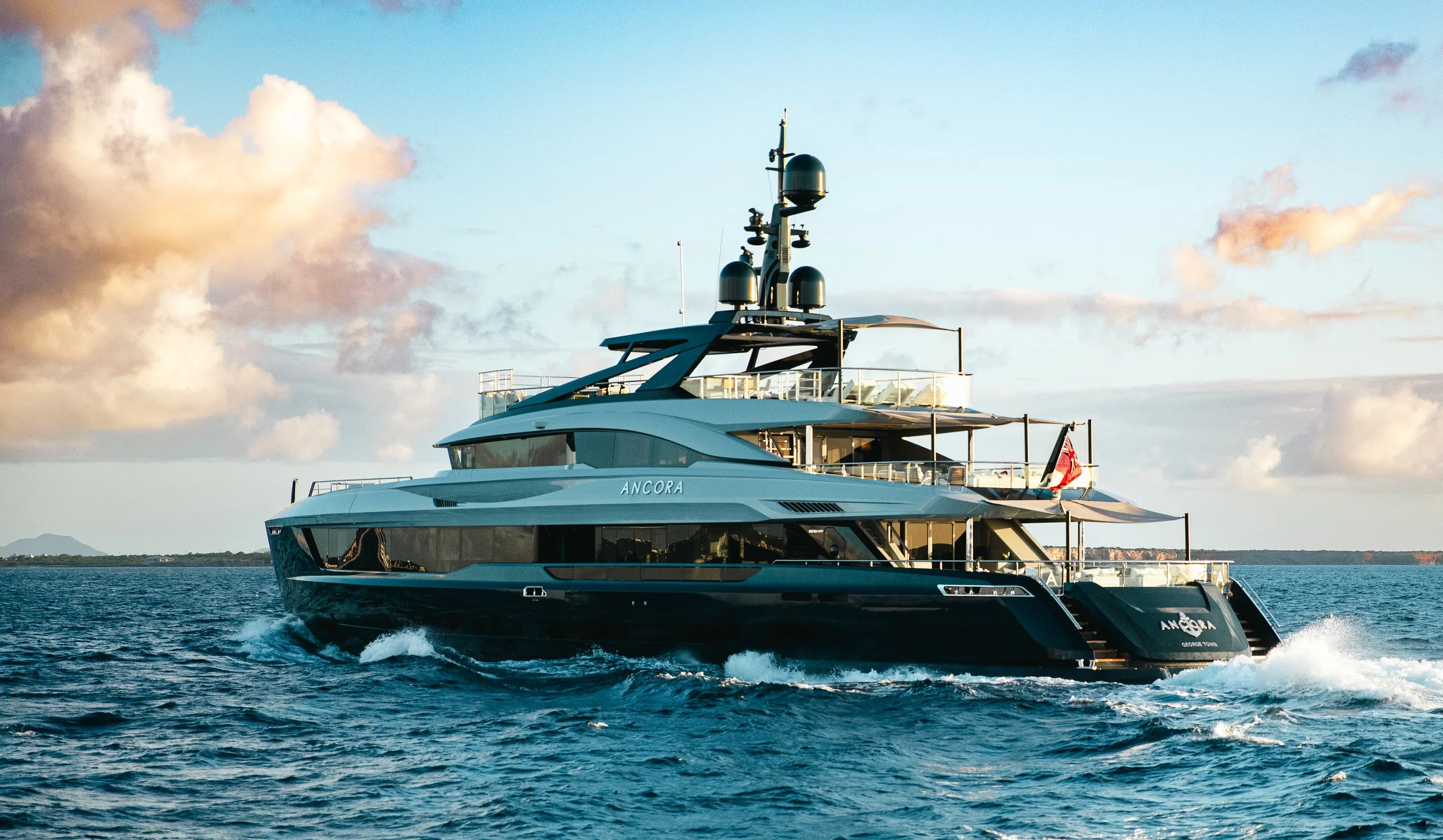 Large luxury yacht named 'ANCORA' sailing on the ocean with a landmass in the distance, under a sky with scattered clouds.