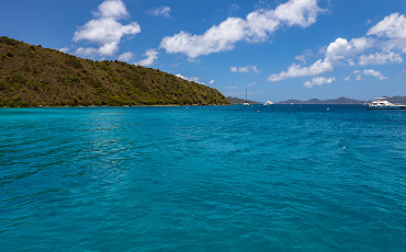 Dark teal water glimmers under a bright partly cloudy sky, next to green land mass.