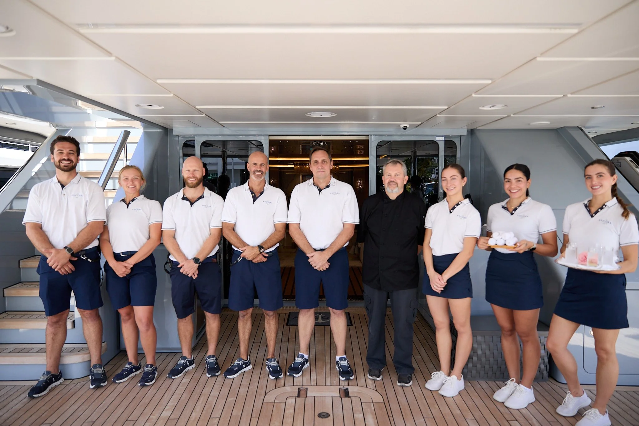 Group of nine yacht staff members standing on the deck of a yacht, dressed in uniform, posing for a photo.
