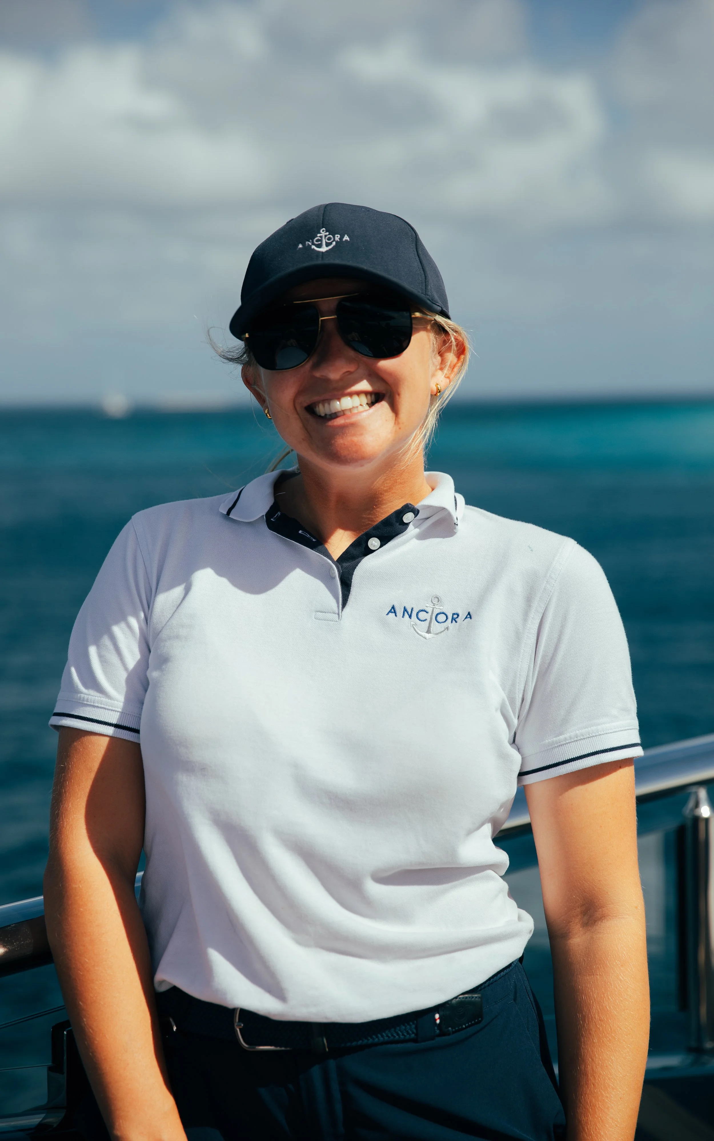A woman smiling on a boat, wearing a white polo shirt with 'ANCHOR' logo, a navy cap, and sunglasses, with the ocean and sky in the background.
