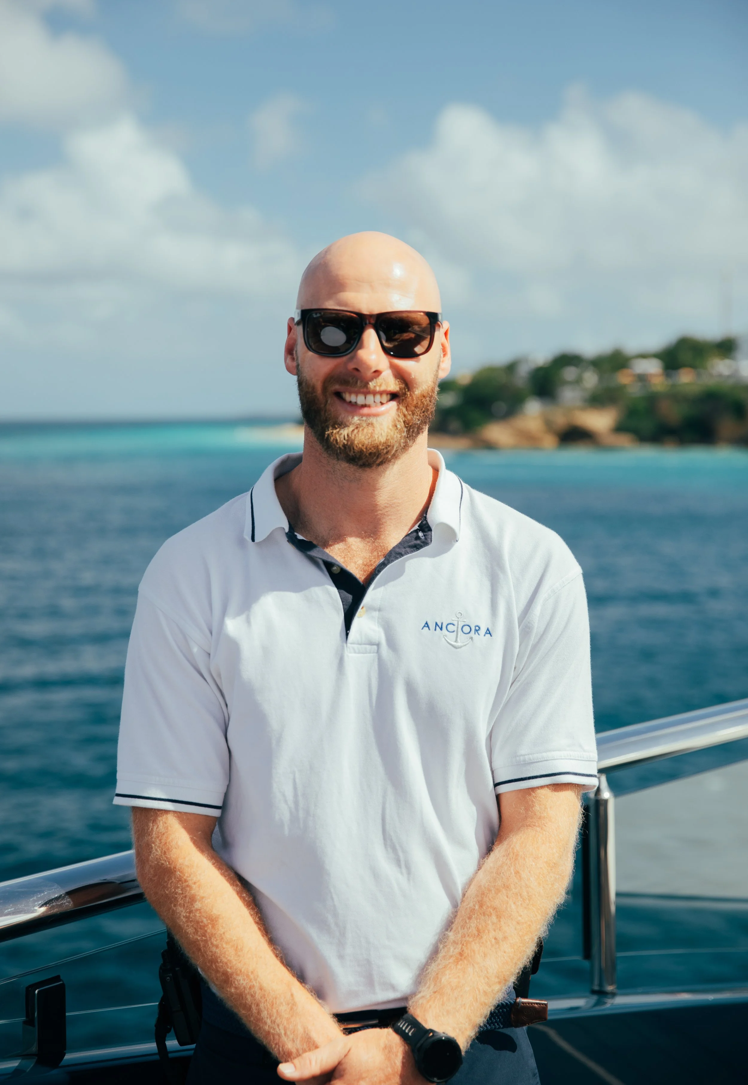 A smiling man with a beard, wearing sunglasses and a white polo shirt with the logo 'ANCORA,' standing on a boat with a body of water and a distant shoreline in the background.