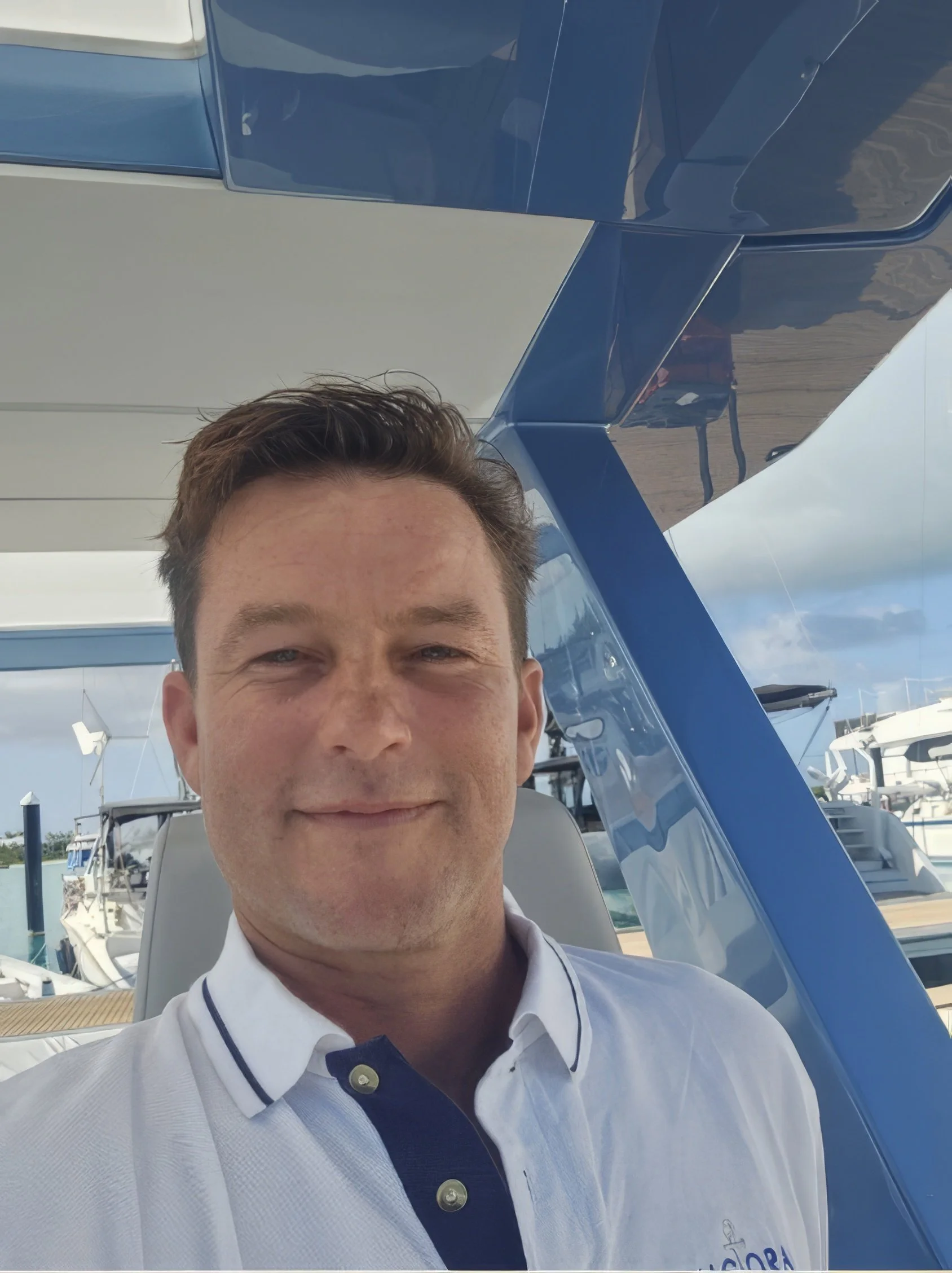 A man smiling on a boat dock with yachts in the background, under a partly cloudy sky.