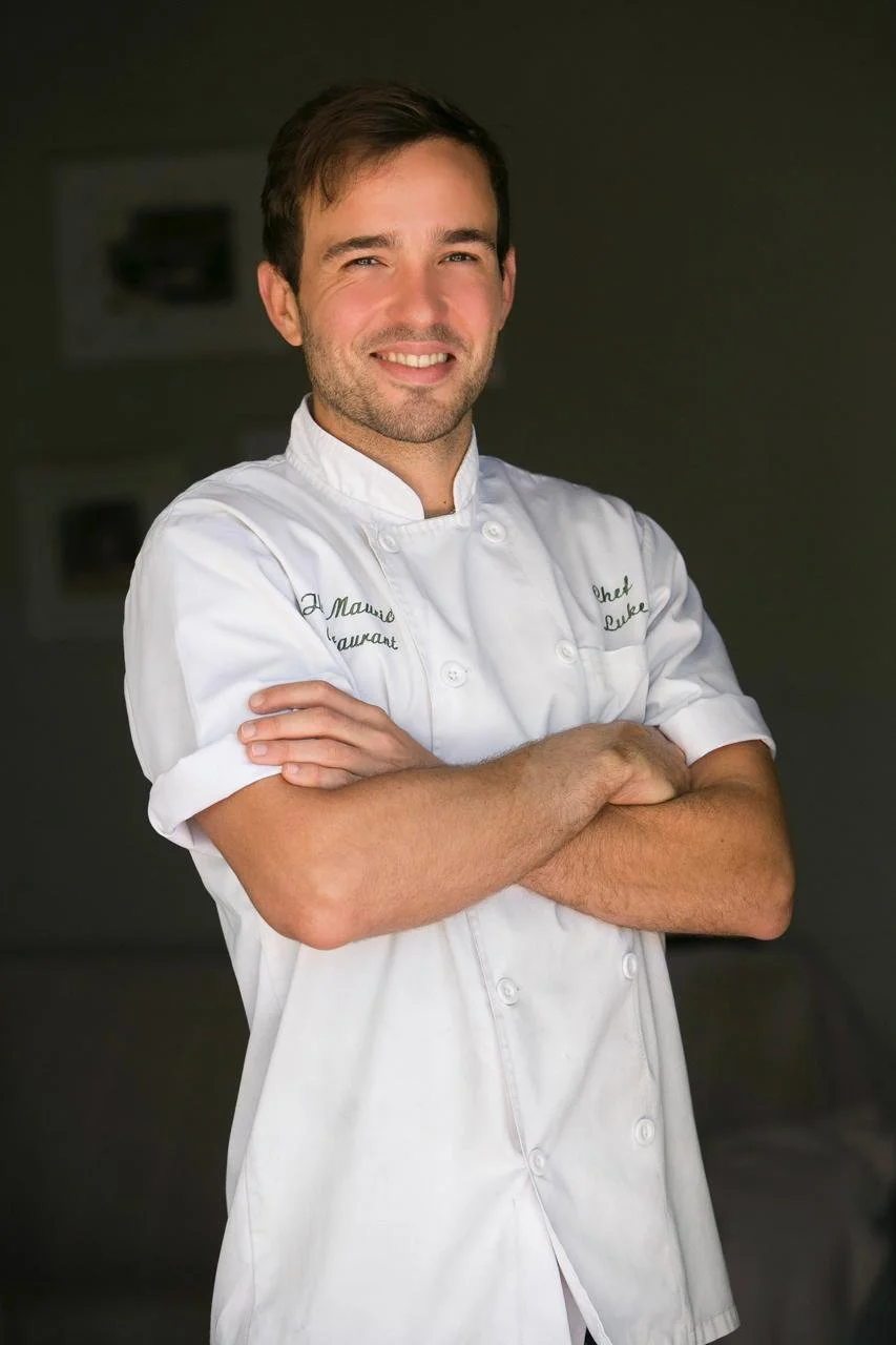Portrait of a male chef in a white uniform with crossed arms, smiling, standing indoors.