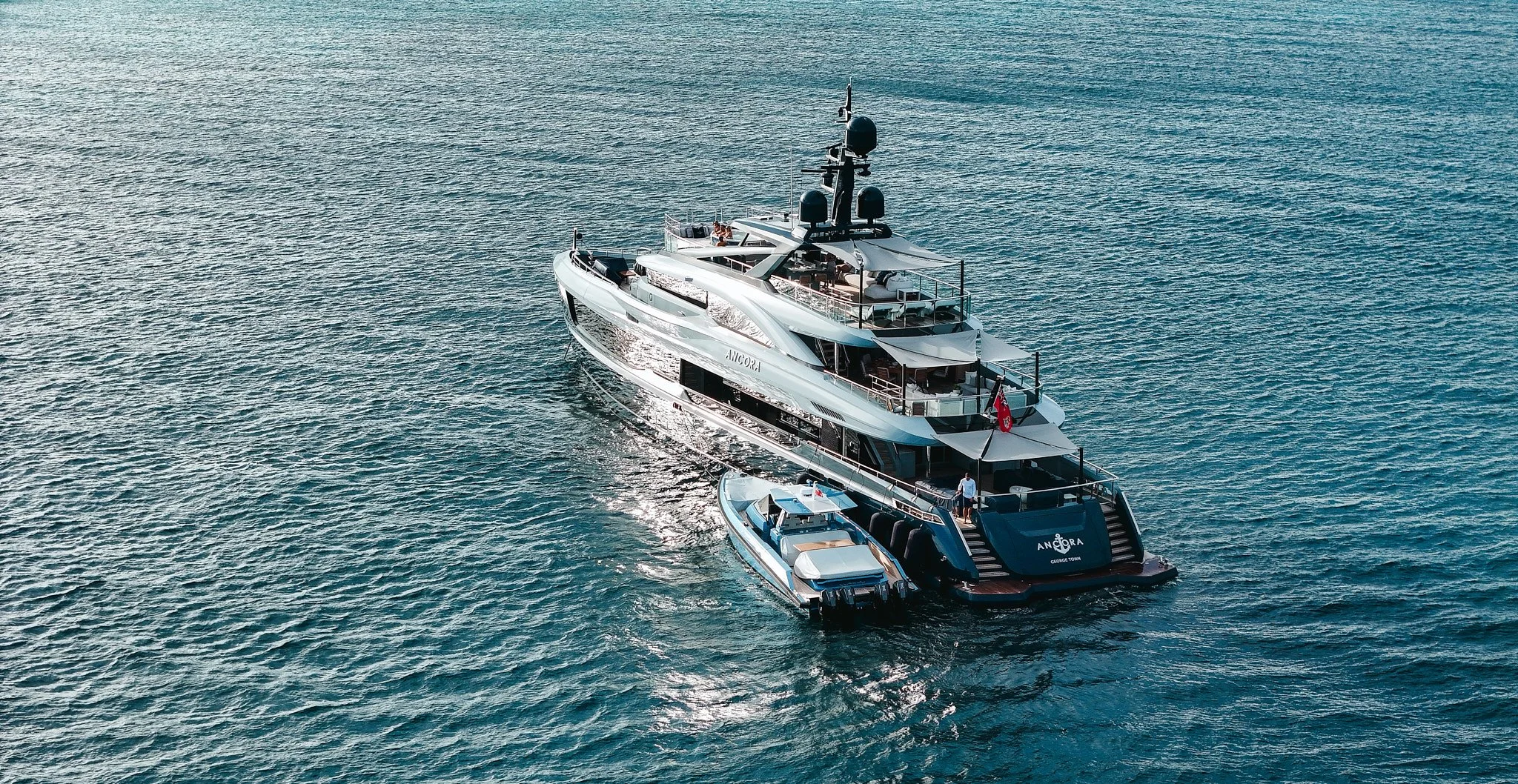 A large luxury yacht with a smaller boat attached to it is sailing on the ocean, with clear skies and calm water.