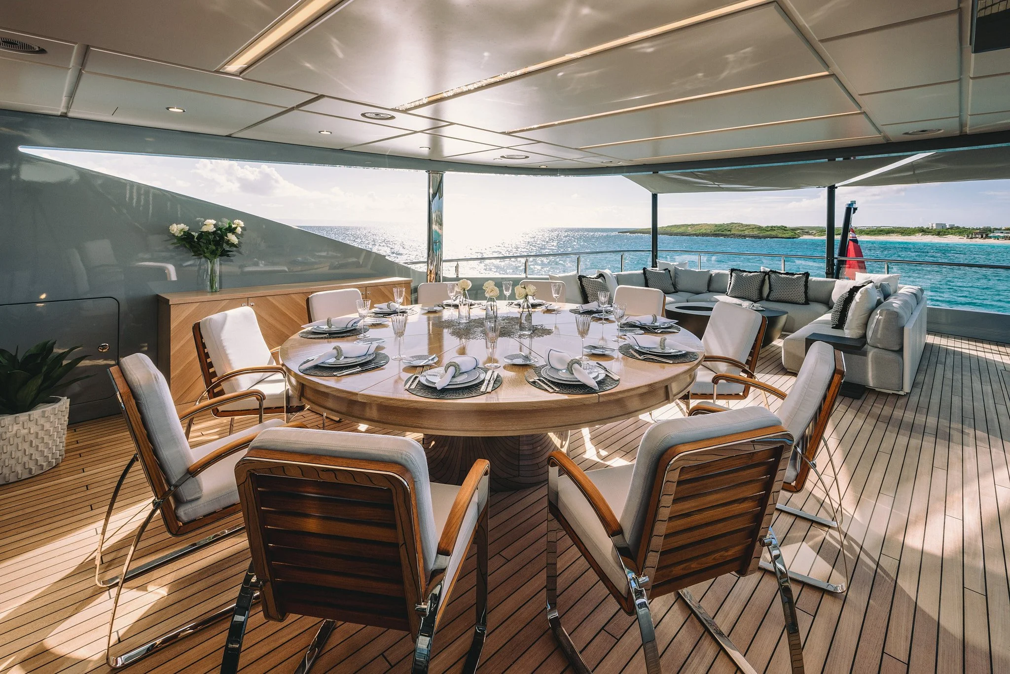 Empty outdoor dining area on a yacht deck with a round table set with plates, glasses, and napkins, overlooking the ocean with a distant landmass.