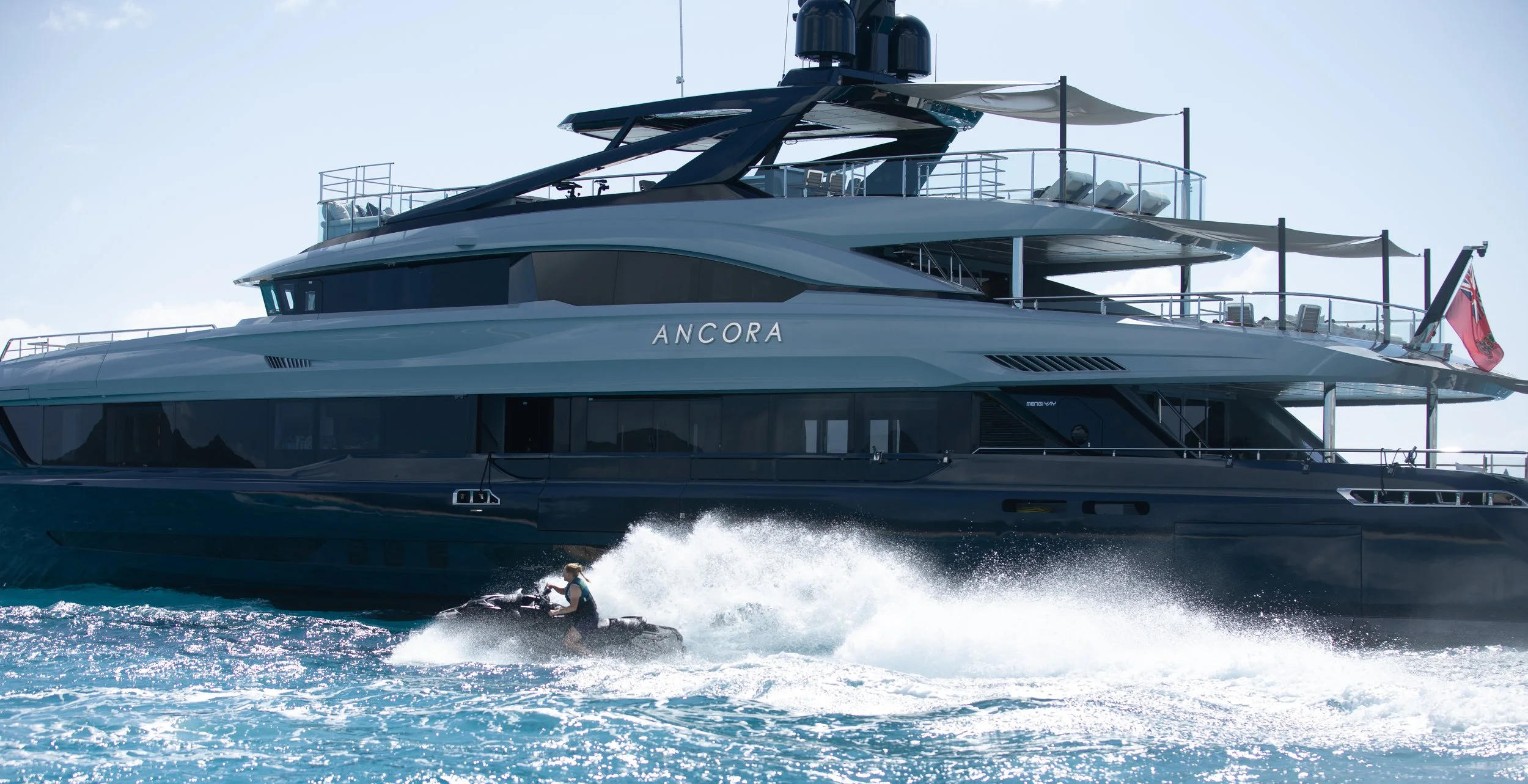 A large, modern luxury yacht named 'Ancora' sailing on the water with a woman riding a jet ski nearby.