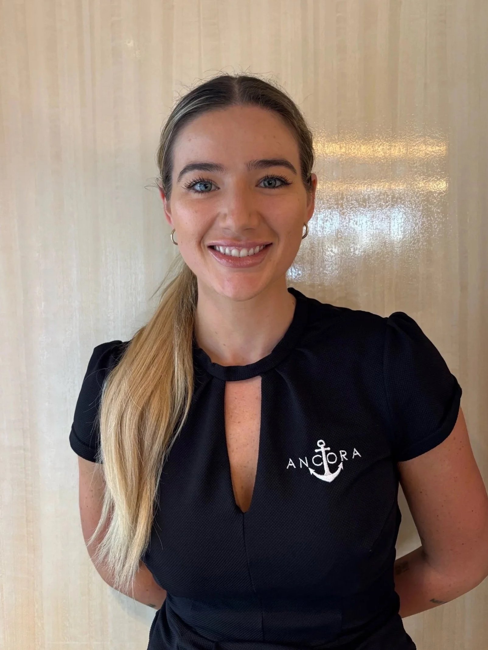 A woman with long blonde hair, blue eyes, and hoop earrings, smiling, wearing a black top with an anchor logo and the word 'Anchora' on it, standing in front of a beige background.