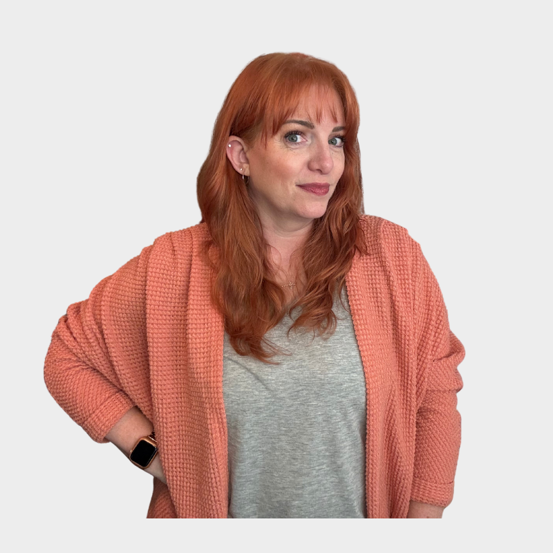 A woman with red hair and blue eyes wearing a pink cardigan over a gray shirt, standing with one hand on her hip against a plain white background.