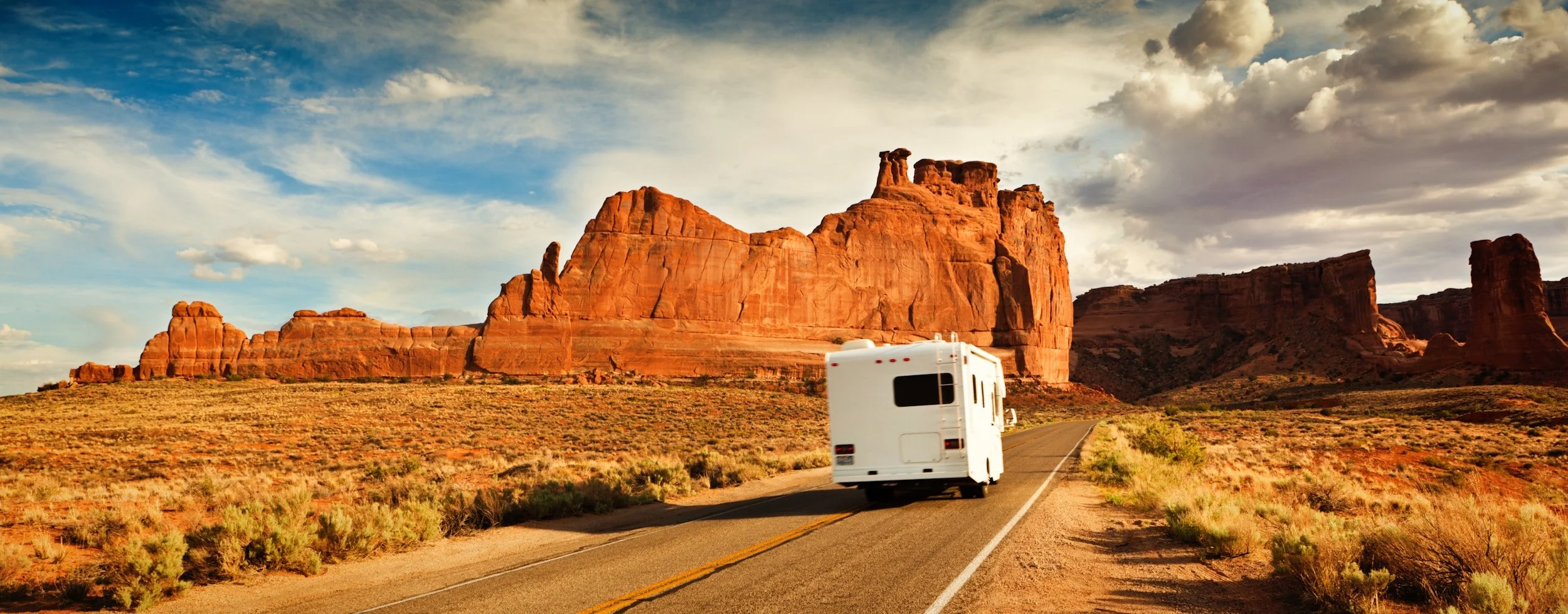 A white RV driving on a desert road with red rock formations and a partly cloudy sky in the background.