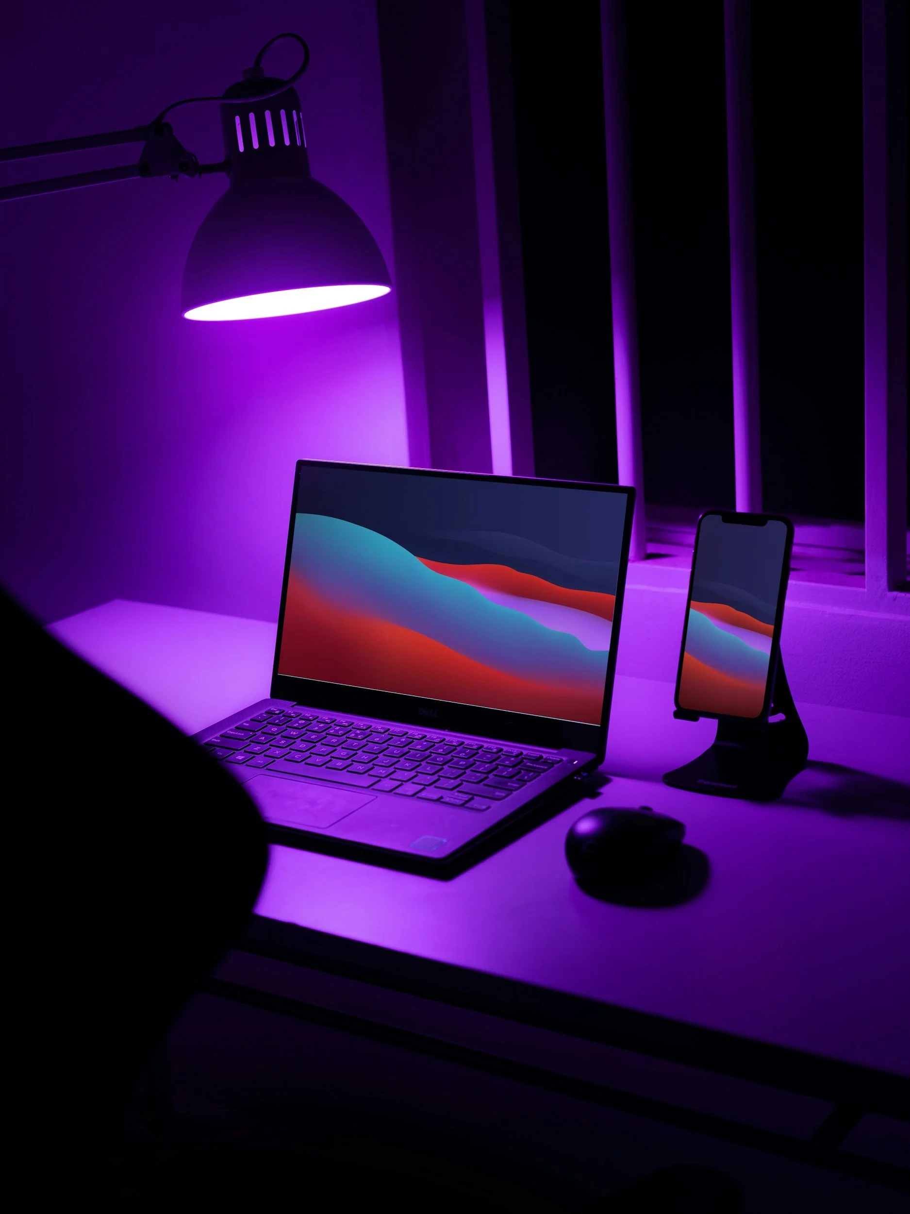 A workspace with a laptop displaying a colorful abstract background, a smartphone in a stand, a computer mouse, and a purple desk lamp illuminating the scene with purple light.