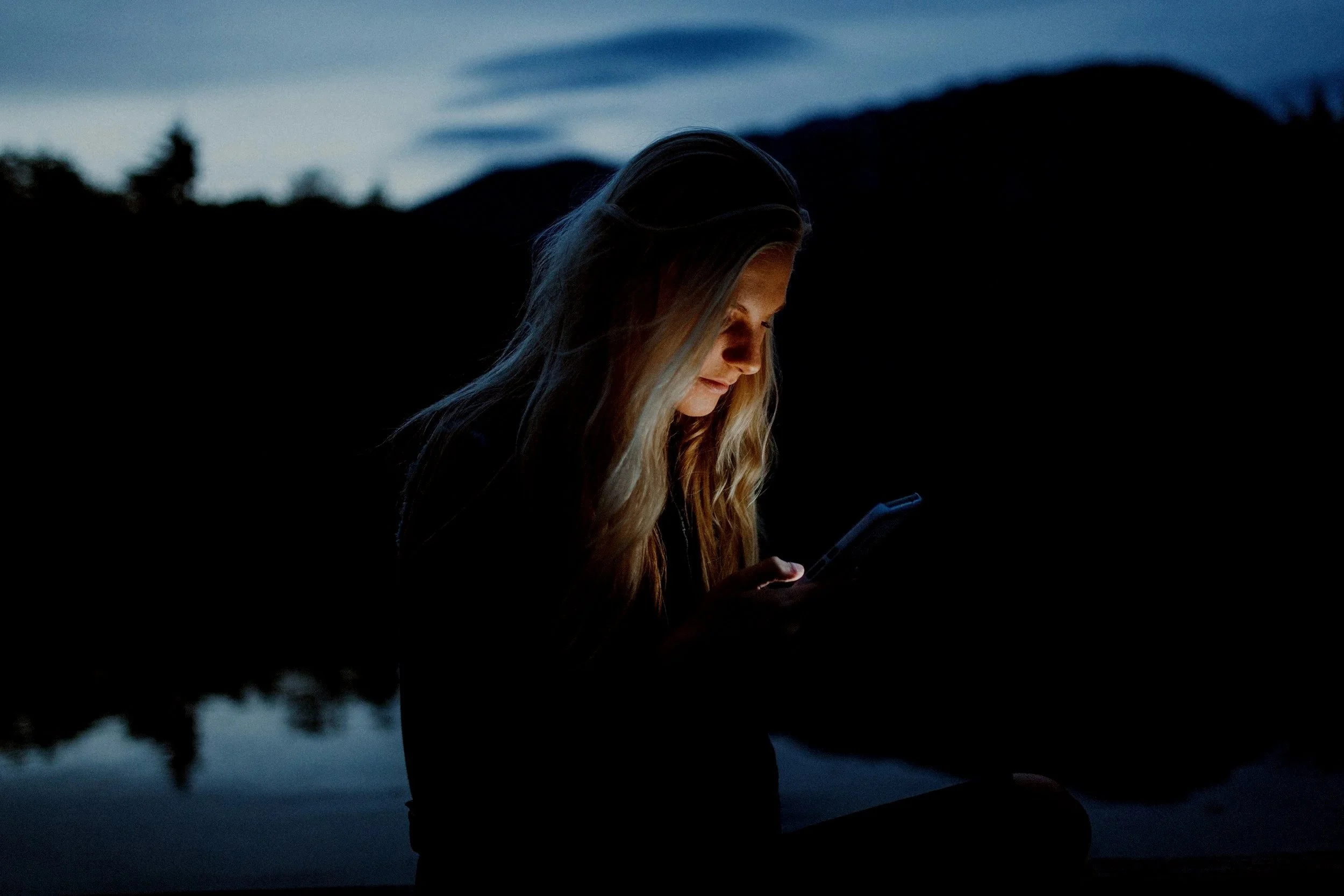 A woman outdoors at night, sitting by a body of water, illuminated by her phone's screen, with dark hills and a cloudy sky in the background.