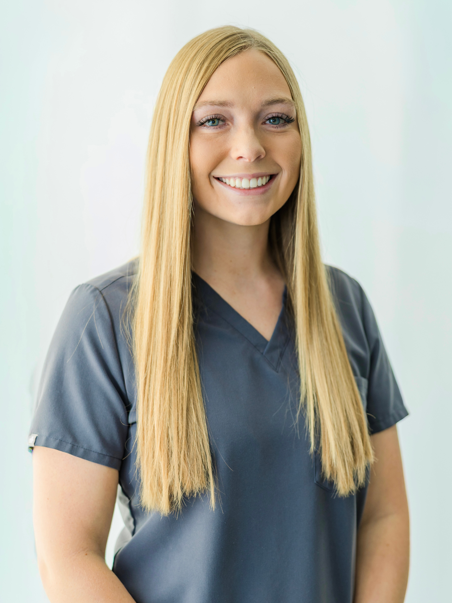 A woman with long blonde hair and blue eyes, smiling, wearing a dark gray scrub top, standing against a plain light background.