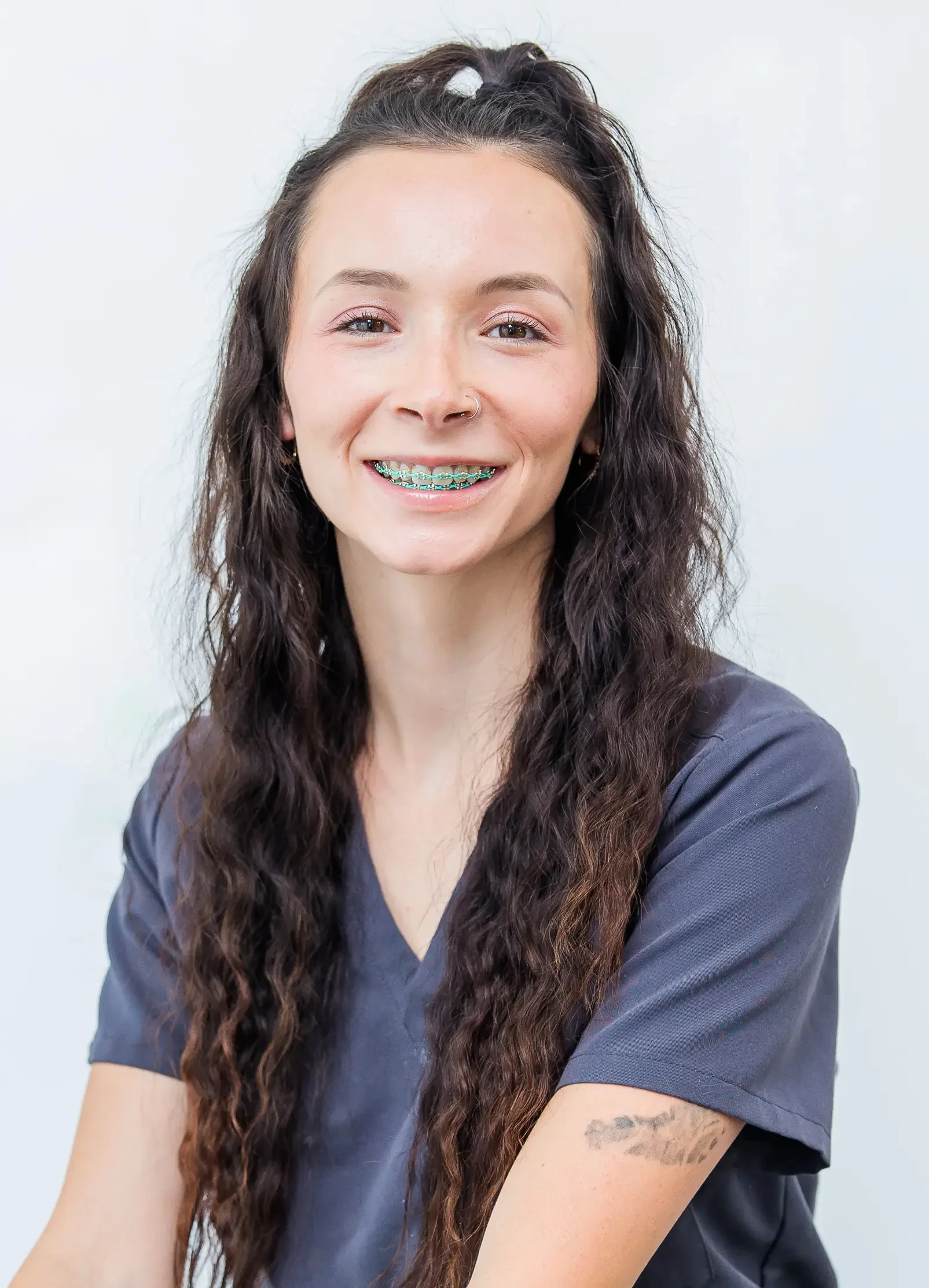 A smiling young woman with long curly brown hair, with teal braces and a gray scrub top, sitting against a plain white background.
