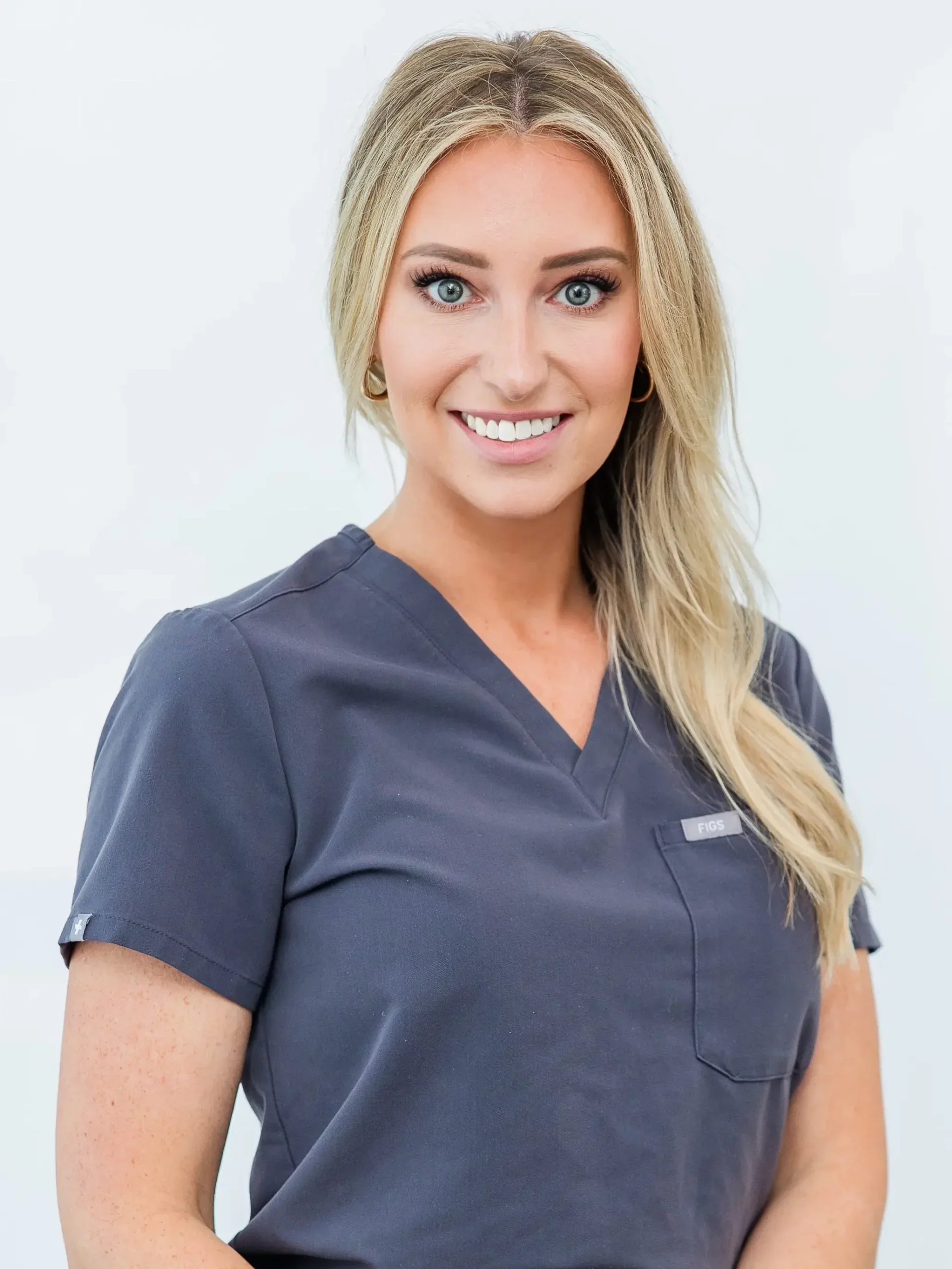 Woman with long blonde hair and blue eyes, wearing a dark gray top, smiling against a plain white background.