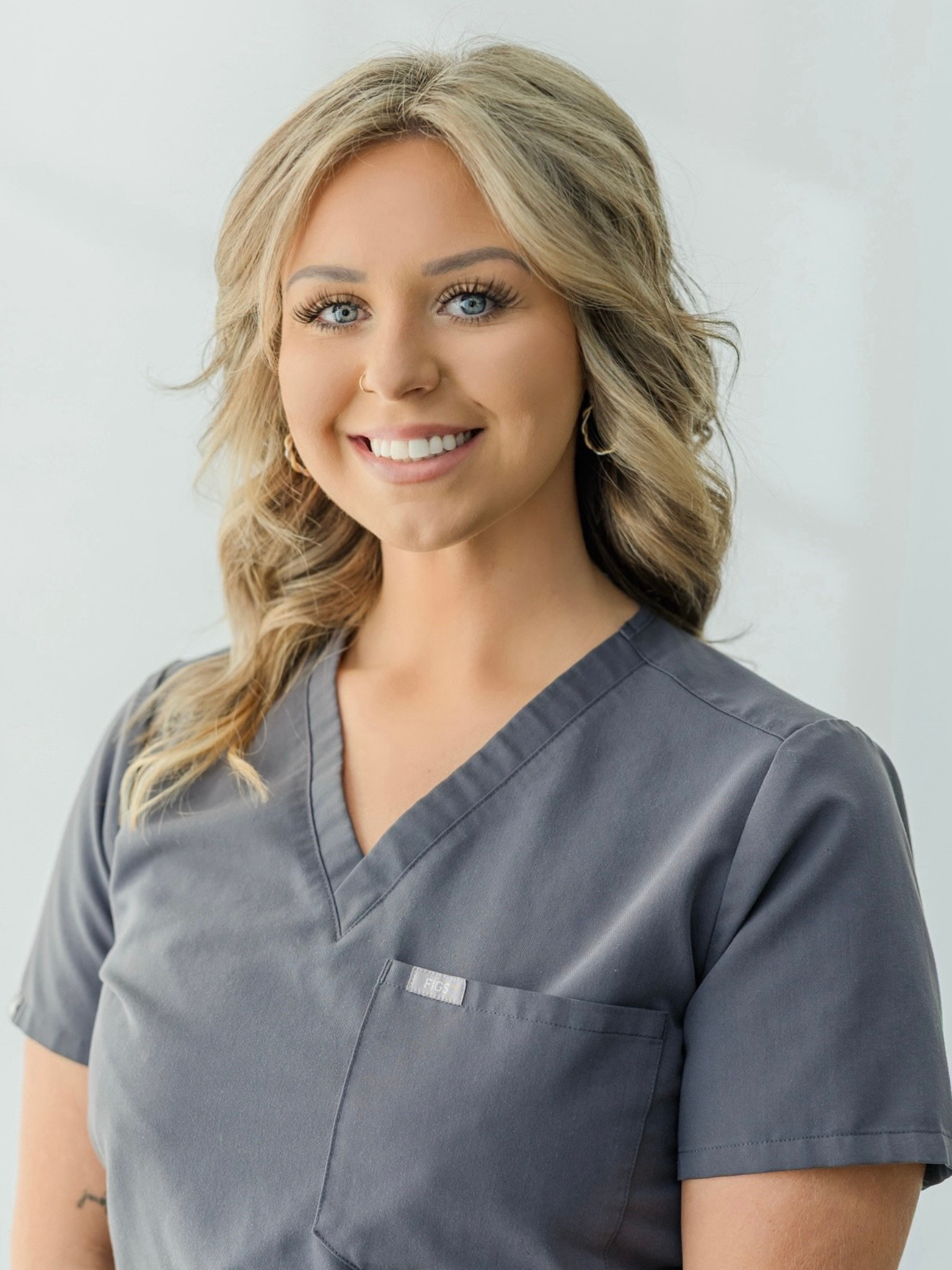 A smiling woman with blonde hair, blue eyes, wearing gray scrubs, standing against a plain light background.
