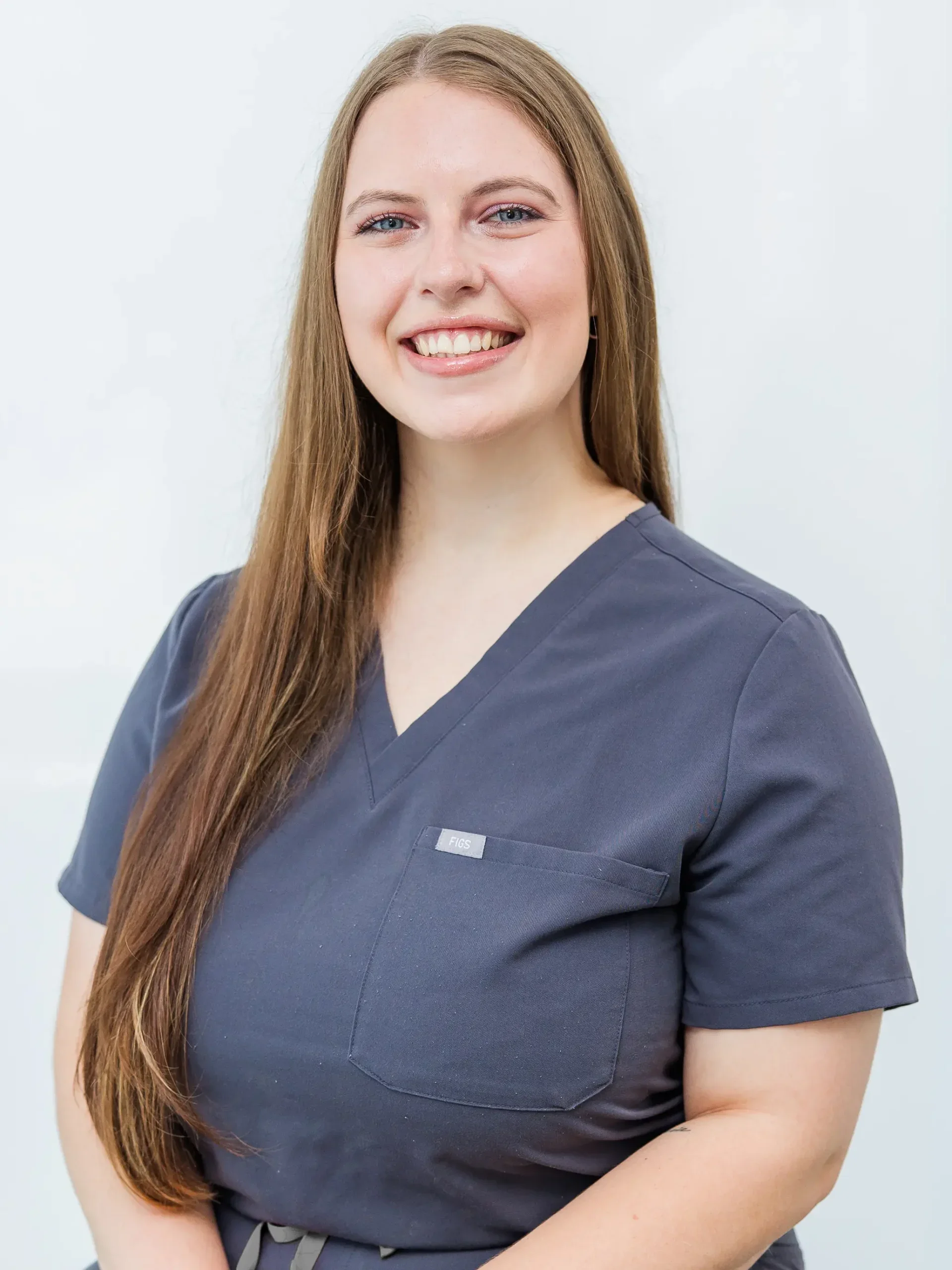 A woman with long brown hair wearing navy blue medical scrubs, smiling against a plain white background.