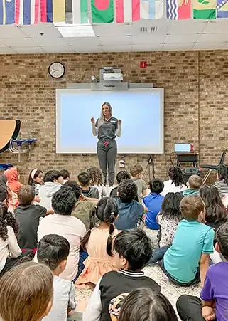 A teacher standing in front of a classroom of young students, giving a presentation. The classroom has a brick wall, a large whiteboard, and a clock. Students are sitting on the floor, facing the teacher.