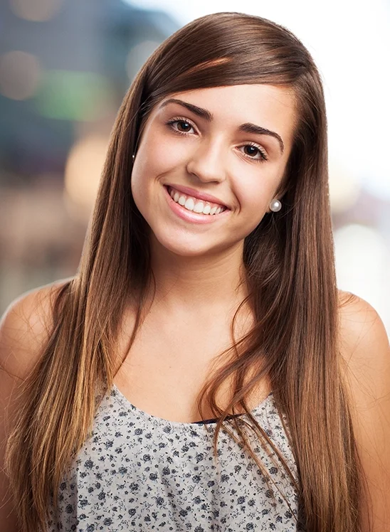 Young woman with long brown hair, wearing a sleeveless floral top and pearl earrings, smiling at the camera.