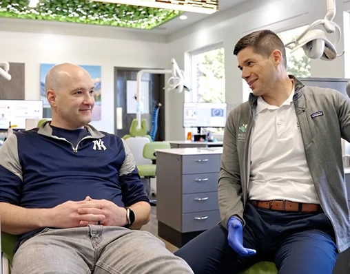 A person in a New York Yankees jacket sitting in a dental or medical office, talking to a healthcare professional with a glove on. The healthcare professional is smiling and making eye contact.