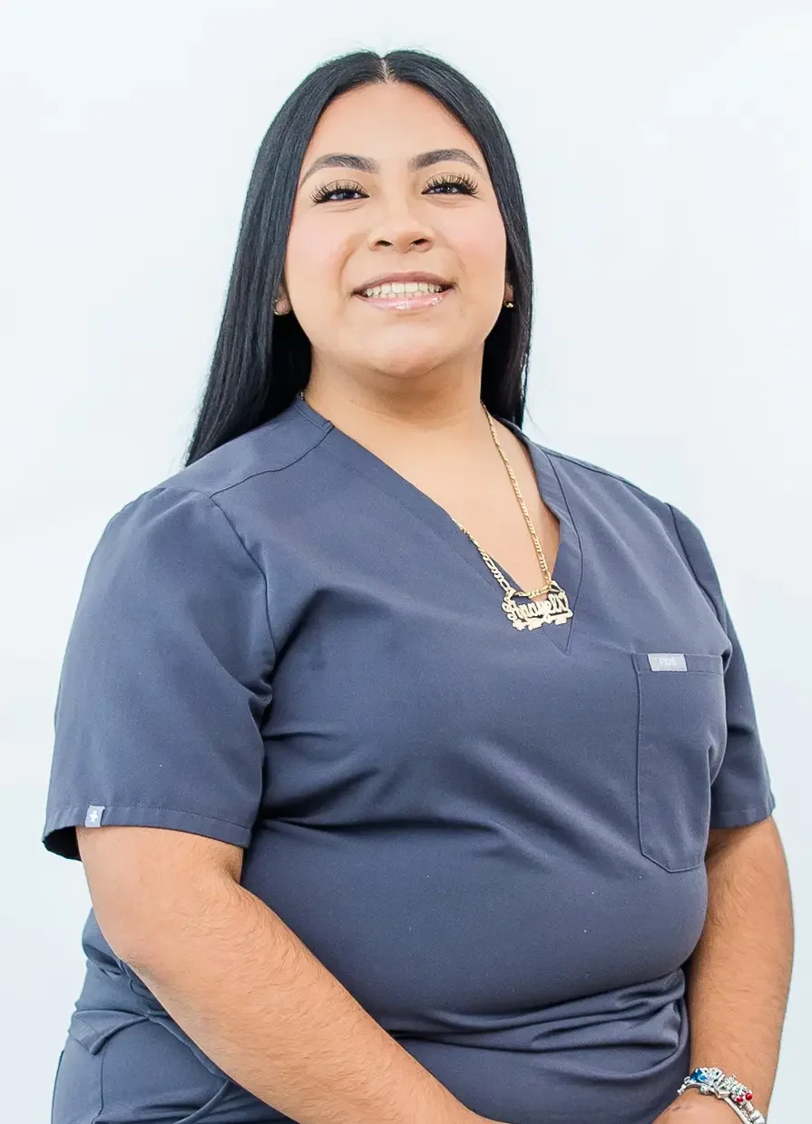 A smiling woman with long black hair wearing navy scrubs and jewelry, standing against a plain white background.