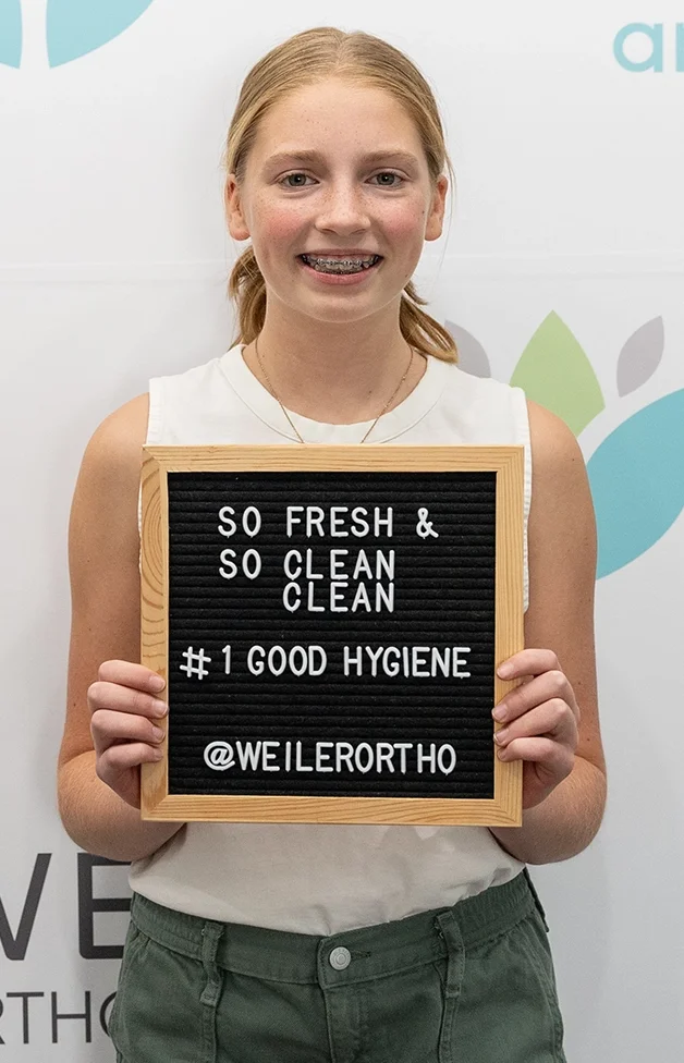 A young girl with red hair, wearing a sleeveless white top and green pants, smiling while holding a black felt letter board with a wooden frame that reads, 'SO FRESH & SO CLEAN CLEAN #1 GOOD HYGIENE @WEILERORTHO'.