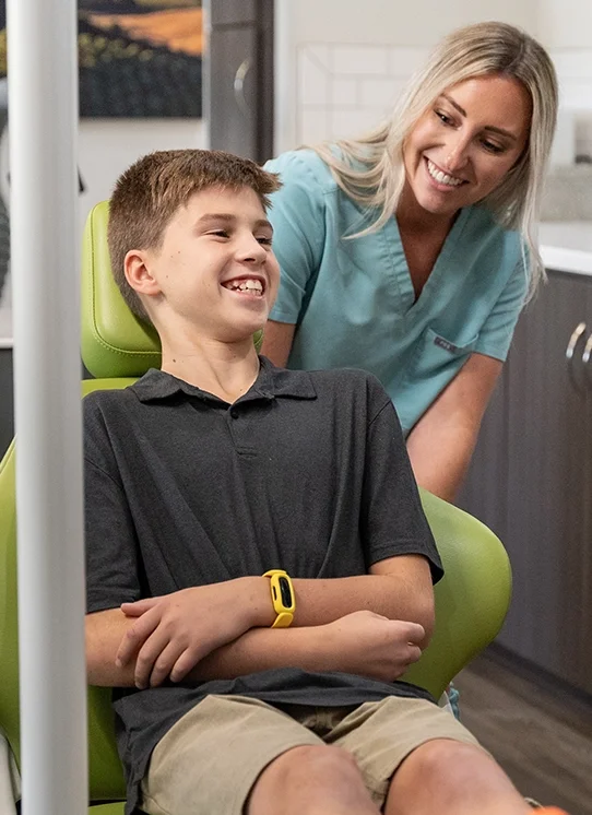 A smiling young boy sitting in a dental chair with a healthcare professional standing beside him in a dental office.