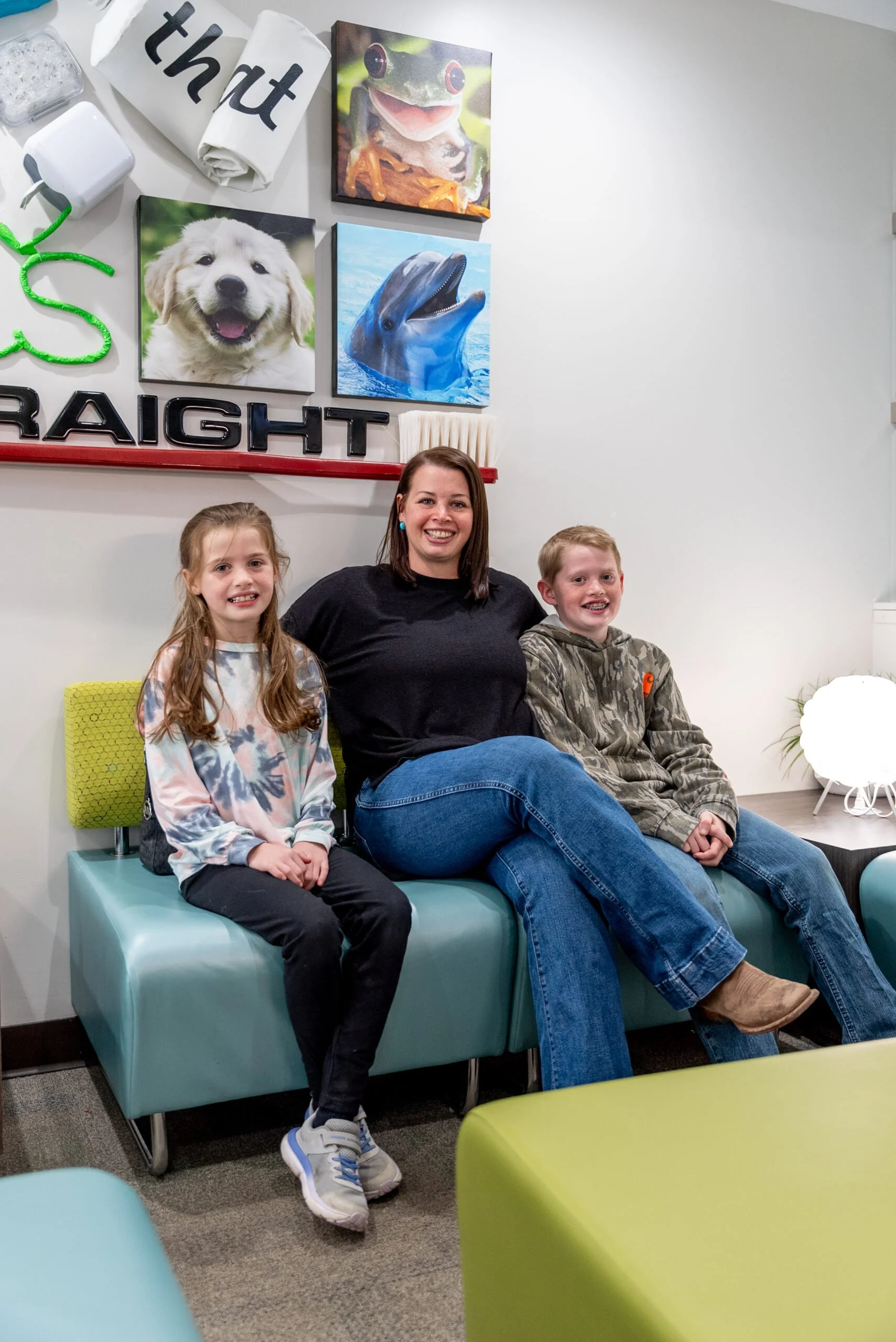 A woman and two children sitting on a teal bench in a room with colorful wall art, including pictures of a puppy, a dolphin, a frog, and a dog. The wall features the words "that" and "straight."