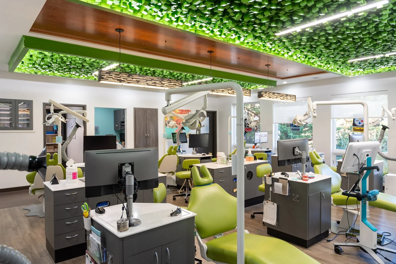 A modern dental clinic with multiple dental chairs, computer workstations, and dental equipment, illuminated by natural light from large windows and decorative green ceiling panels.