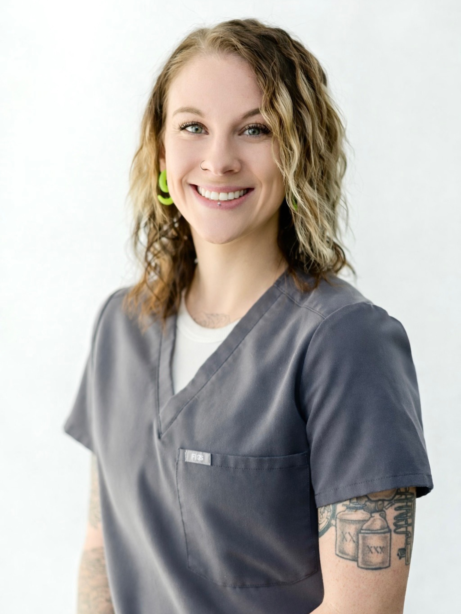 Portrait of a young woman with blonde, curly hair wearing green earrings and gray medical scrubs, smiling at the camera.