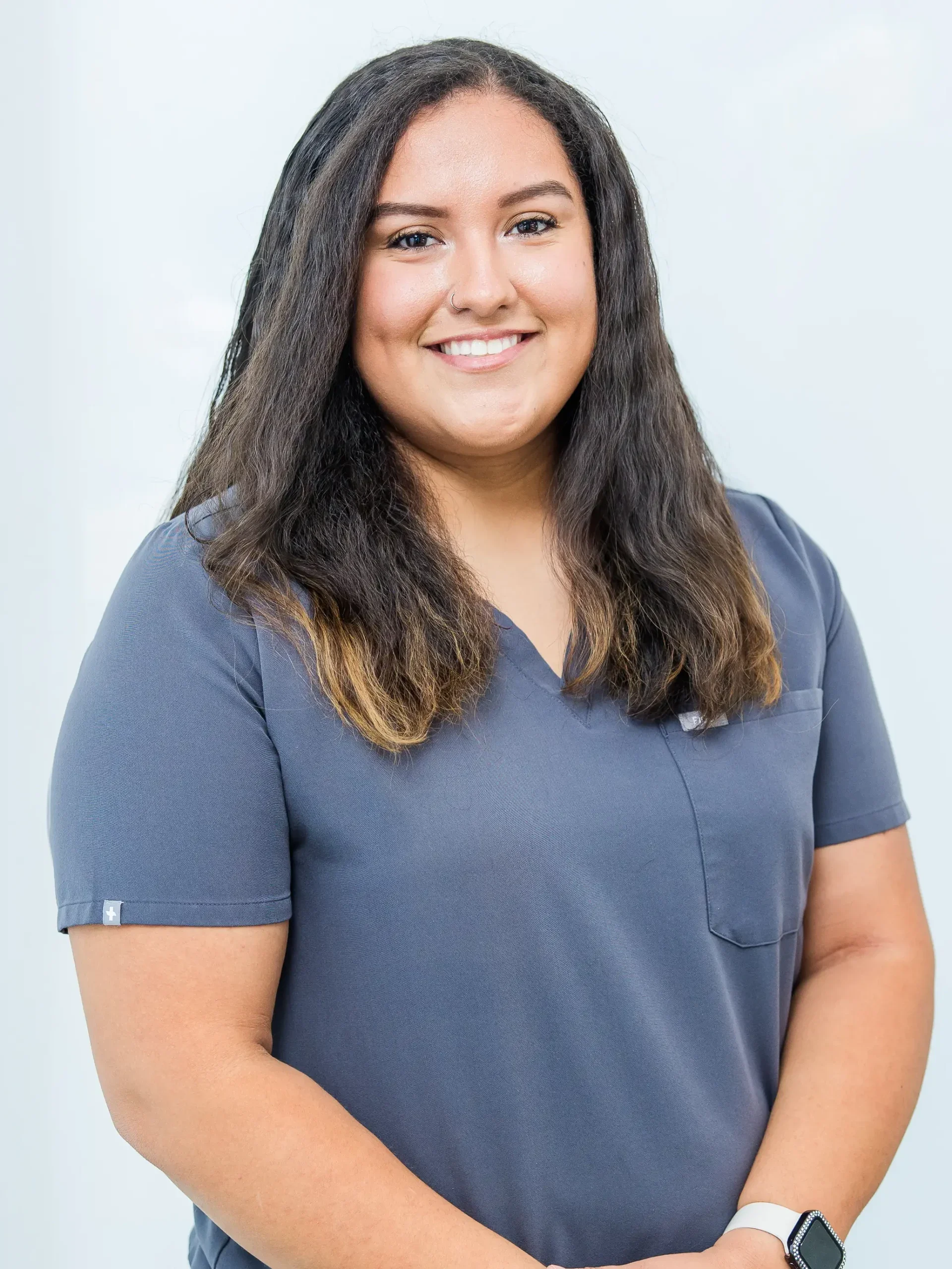 A woman with long dark hair, wearing a navy medical scrub top, smiling, against a light background.