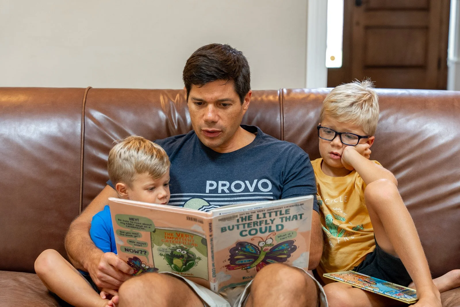 A man reading a colorful children's book called 'The Little Butterfly That Could' to two young boys sitting on a brown leather couch.