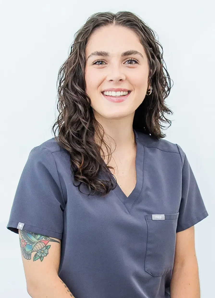 A smiling woman with curly brown hair wearing a navy blue medical scrub top and a tattoo on her left arm, standing against a plain light background.