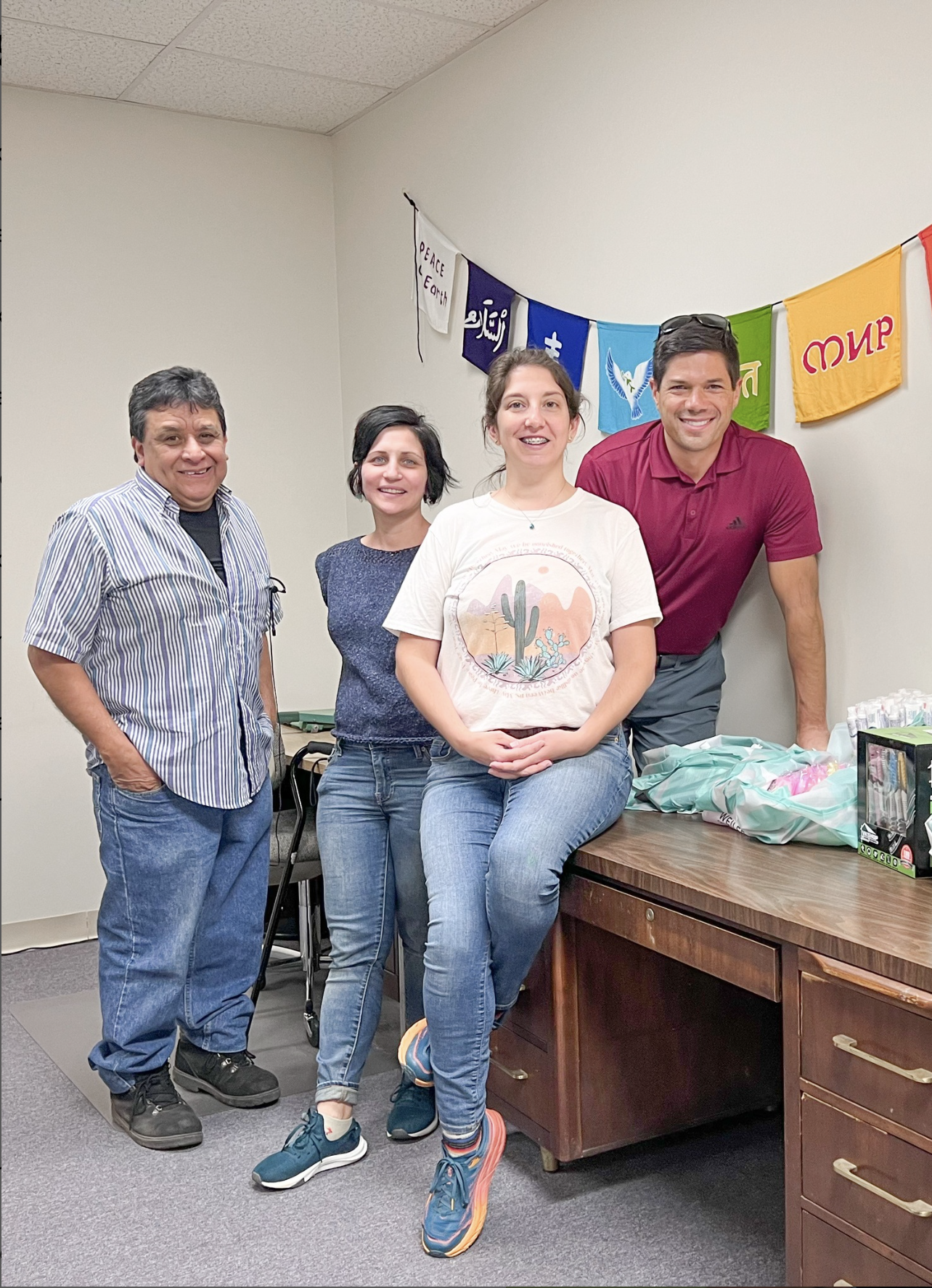 Four people standing and sitting in an office or meeting room, smiling at the camera, with colorful flags hanging on the wall behind them.