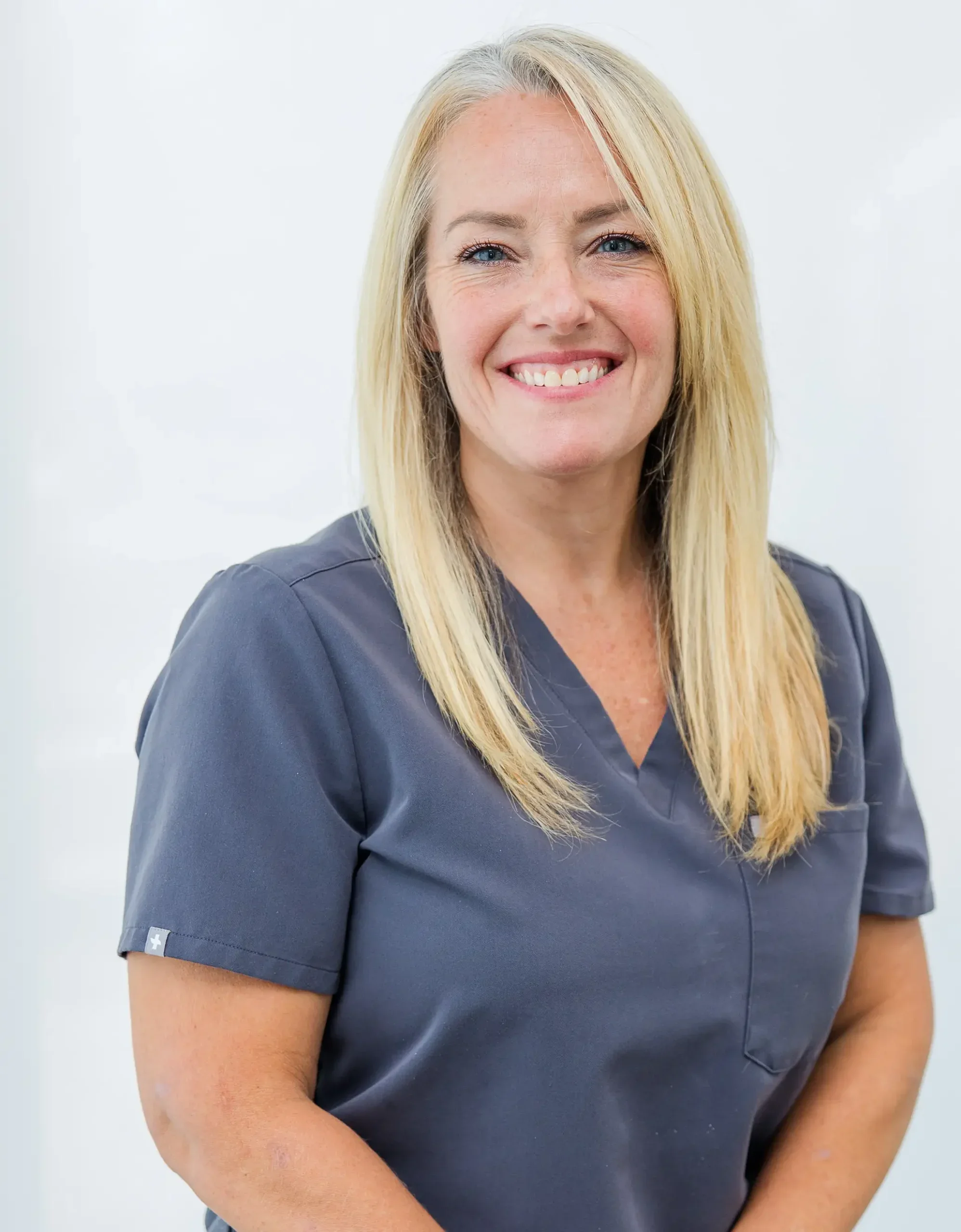 A smiling woman with blonde hair wearing navy blue medical scrubs, standing against a plain white background.