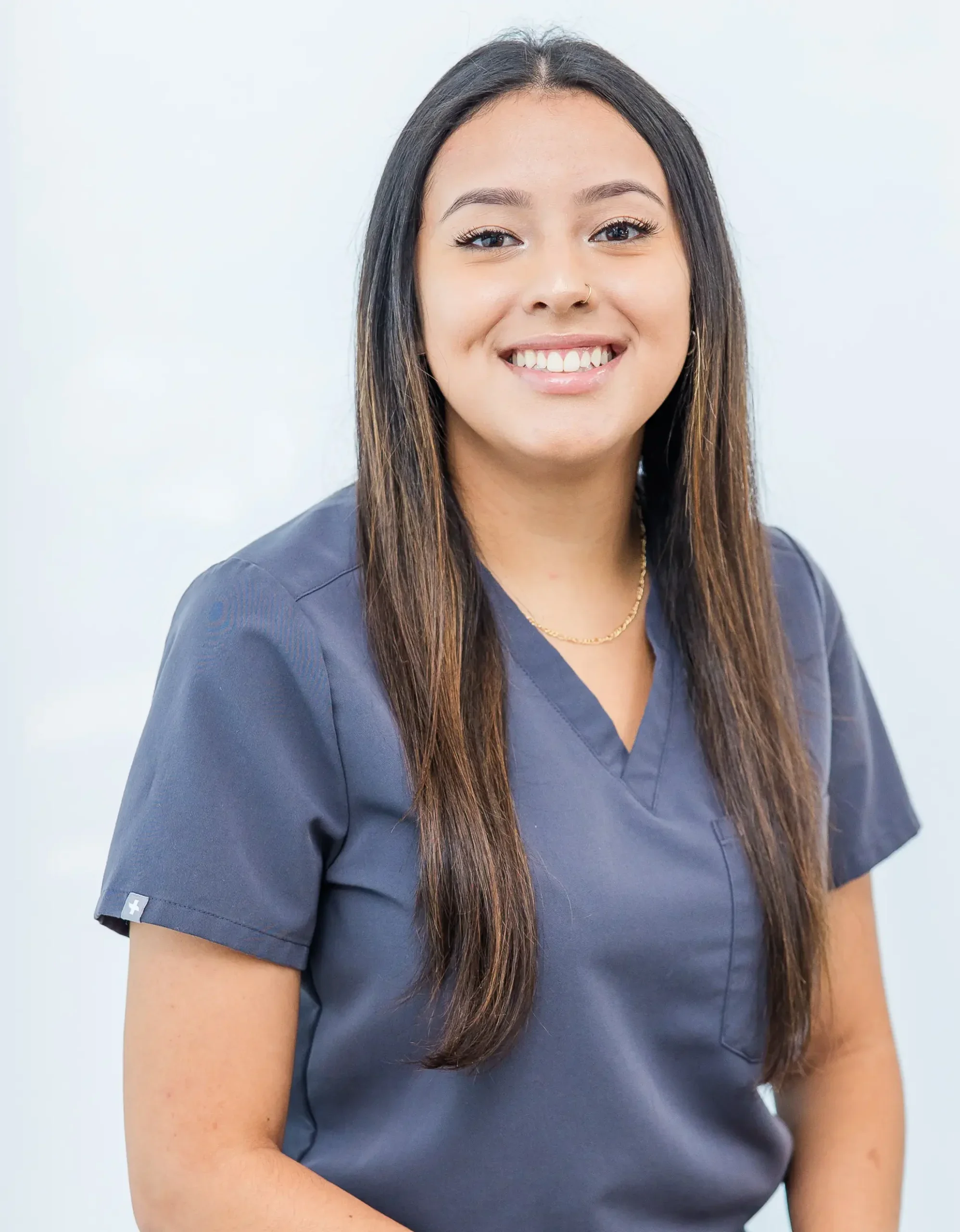 A woman with long dark hair, wearing a gray medical scrub top, smiling, standing in front of a plain white background.