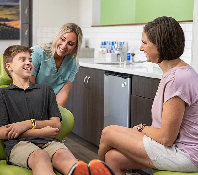 A dentist's office with a young boy sitting in a dental chair, smiling and talking to a female dentist and an adult female, both smiling.