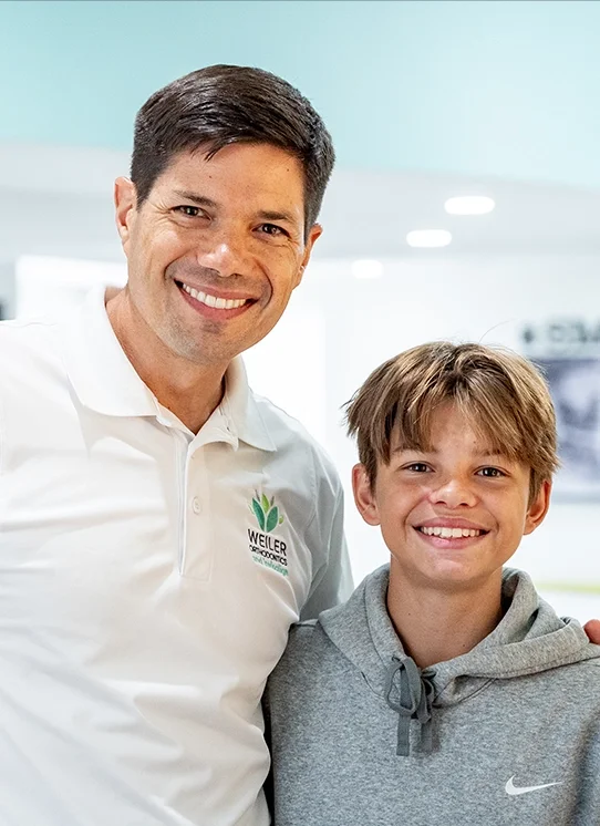 A smiling man in a white polo shirt with a logo standing next to a smiling boy in a gray hoodie, indoors with a light blue wall in the background.