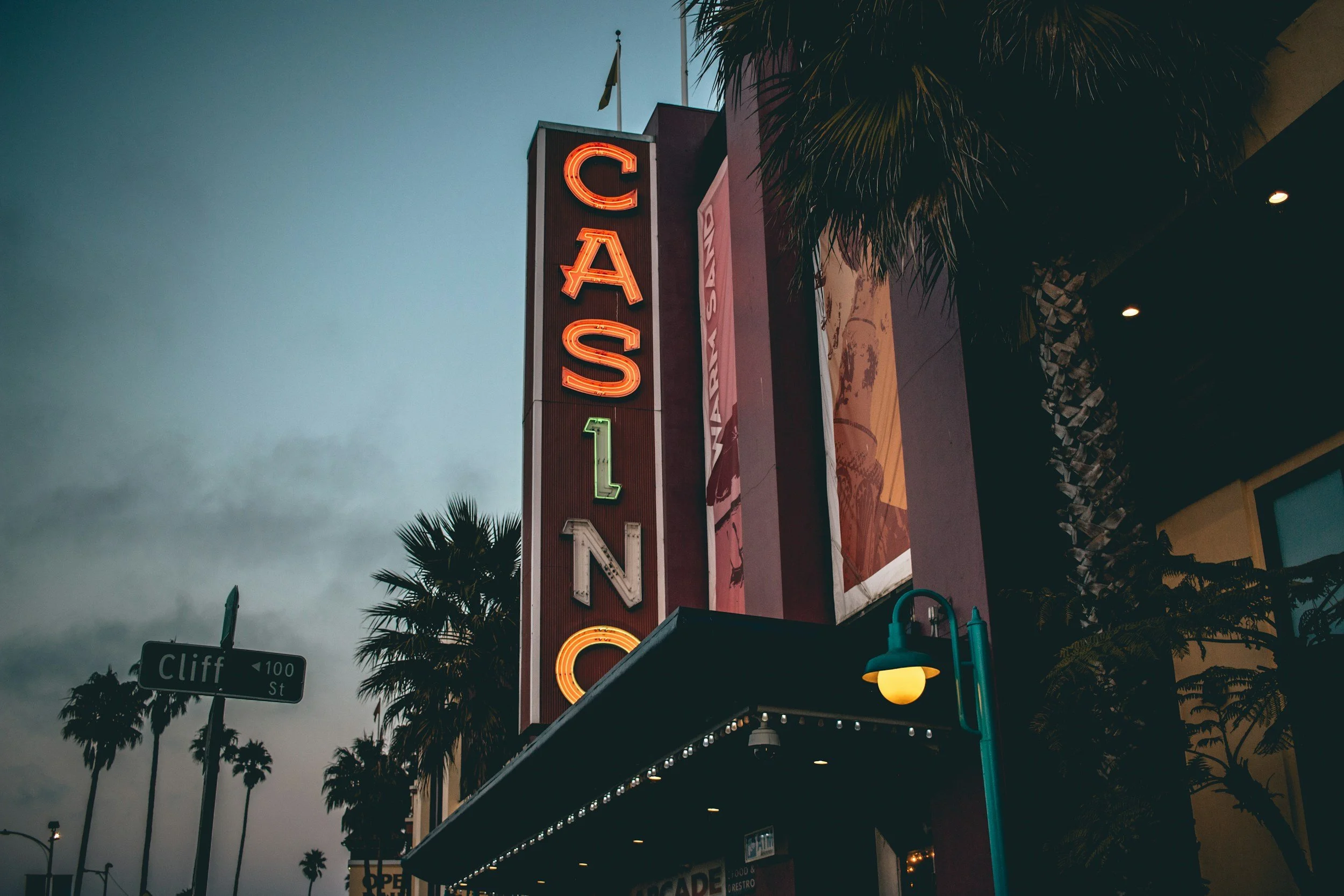The image shows a tall vertical sign with the word 'CASINO' spelled in neon lights. The neighborhood street sign reads 'Cliff 100 St.' Palm trees are visible around the building, and the sky appears to be at dusk or early evening.