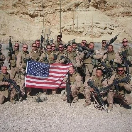 A group of soldiers in desert camouflage uniforms holding firearms. They are standing and kneeling on a gravel surface in front of a rocky, beige background. One soldier is holding an American flag.